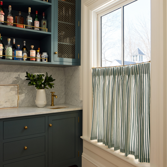 A stylish home bar area features dark teal cabinetry with brass hardware, a marble countertop and backsplash, and a window dressed with pleated green-and-white striped café curtains.