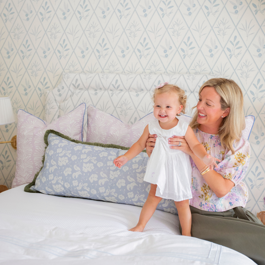 Blonde mother holding her blonde child on white bed styled with three Lavender Botanical Stripe Printed Pillows and one blue floral printed lumbar with sage fringe