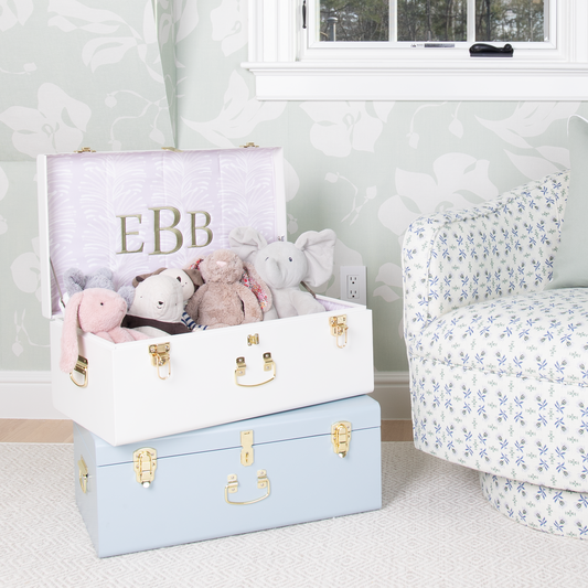Two white and blue keepsake trunks stacked on top of each other in a room with mint floral wallpaper and and a blue and green floral patterned swivel chair