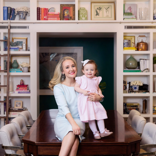 Woman in a blue dress sitting on a wooden table holding a young child in a pink dress in front of a bookcase