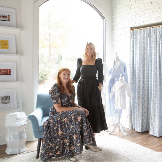 Red headed woman sitting on a blue chair next to a blonde woman standing in a black dress next to two mannequins with robes on in front of a dressing room with sky blue botanical curtains 