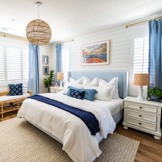 Coastal bedroom with white shiplap walls, blue striped headboard styled with white pillows and sheets, three windows with sky blue velvet curtains hanging on a gold rod