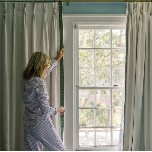 Blonde woman in pajamas closing natural white custom curtains in front of a window 