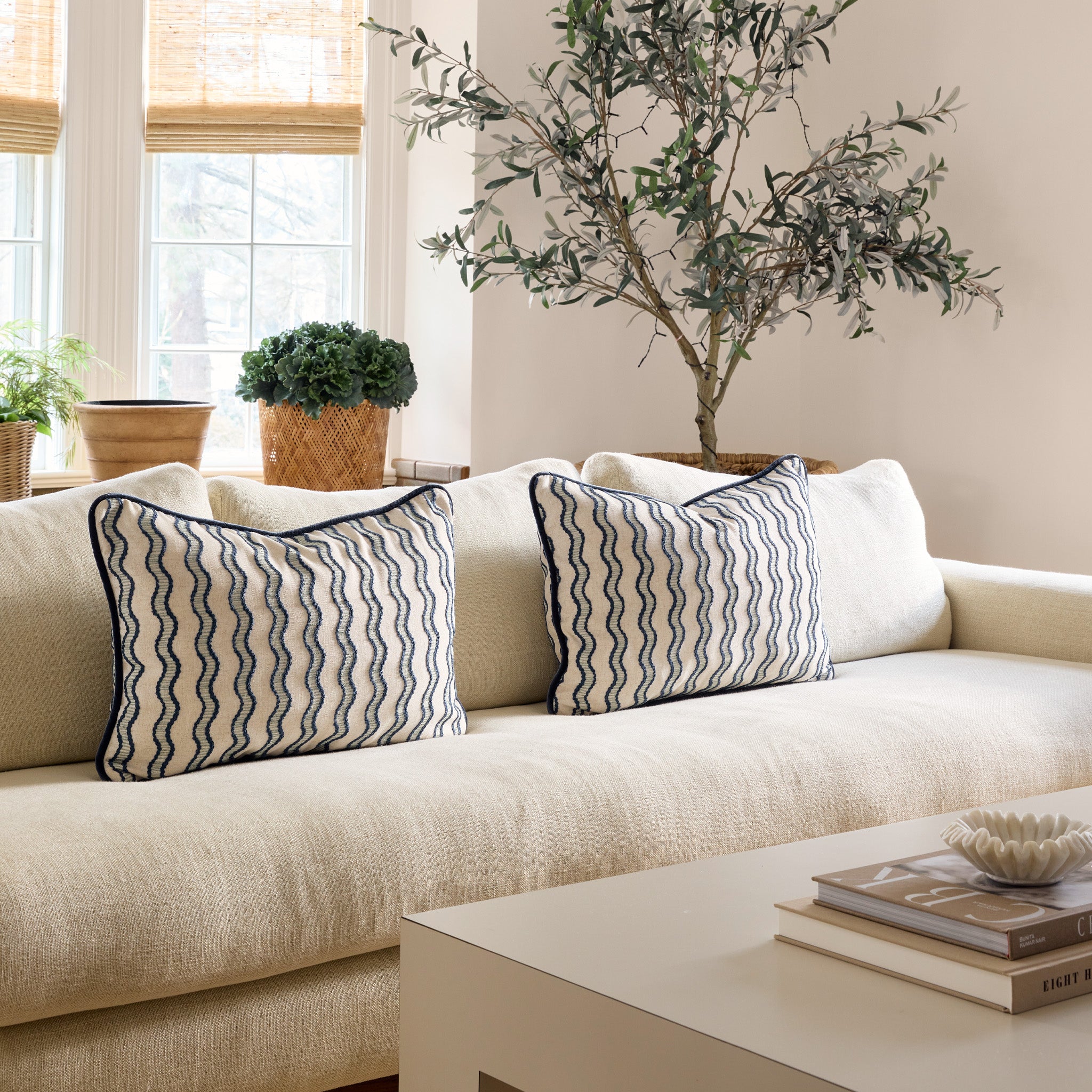 white couch with embroidered cream and navy blue wavy lines pillows on it and illuminated windows behind the couch and a coffee table stacked with books