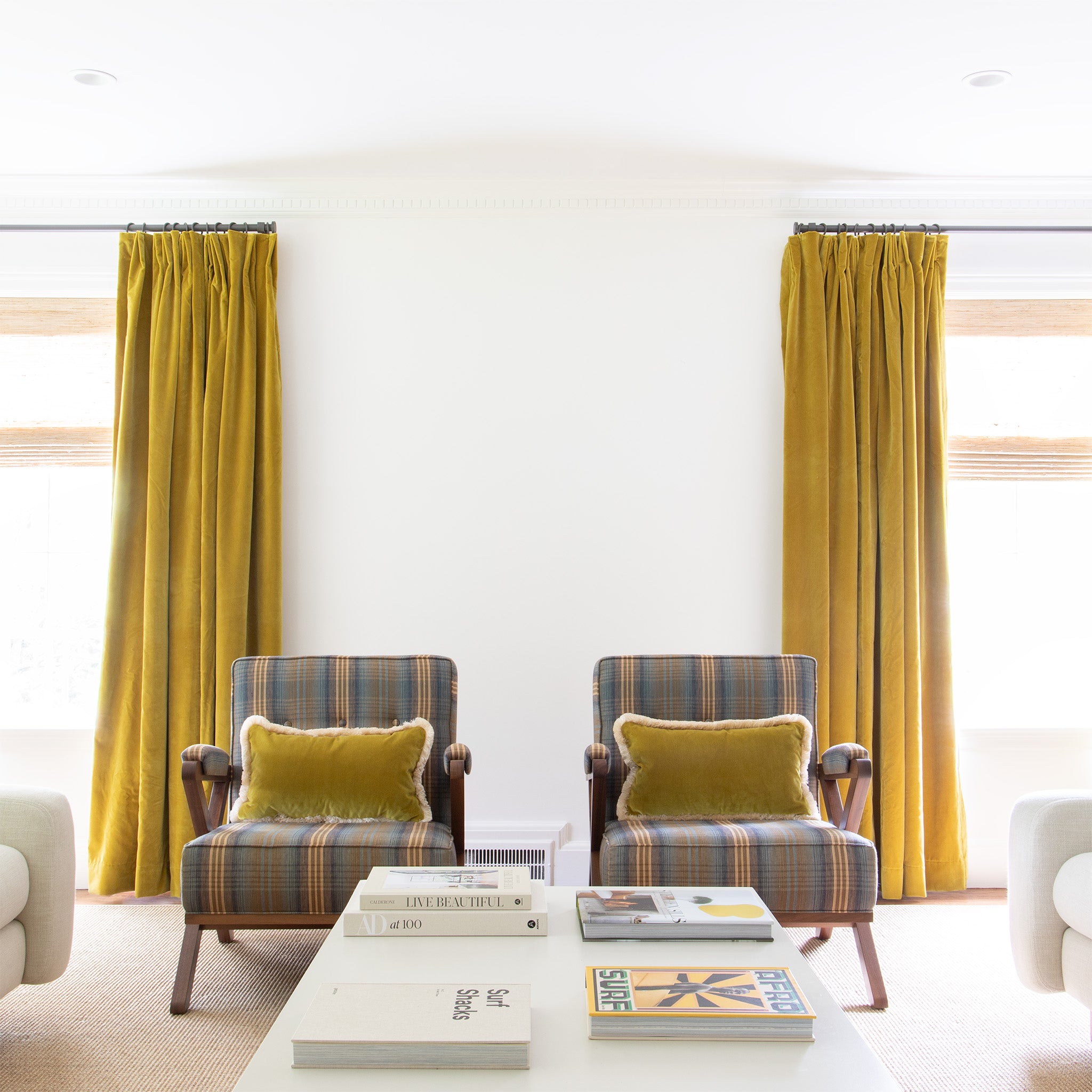 Golden chartreuse velvet curtains on metal rods in front of illuminated windows with blue and yellow striped chairs with Golden chartreuse velvet pillows on them and a white coffee table with books spread on it
