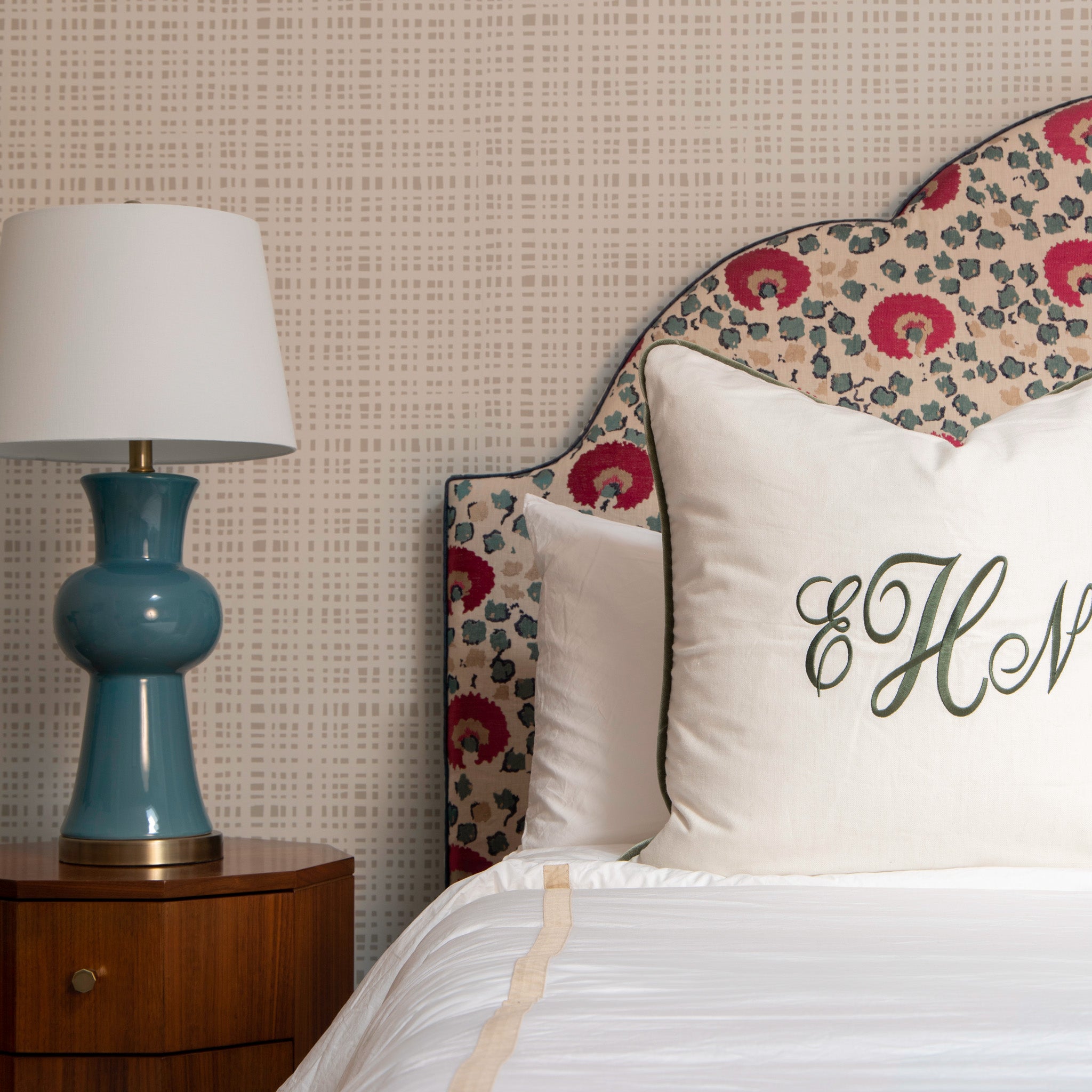 Bed close-up with wooden dresser with blue and white lamp on top by white bed with a natural white linen monogrammed pillow styled with cream gingham printed wallpaper