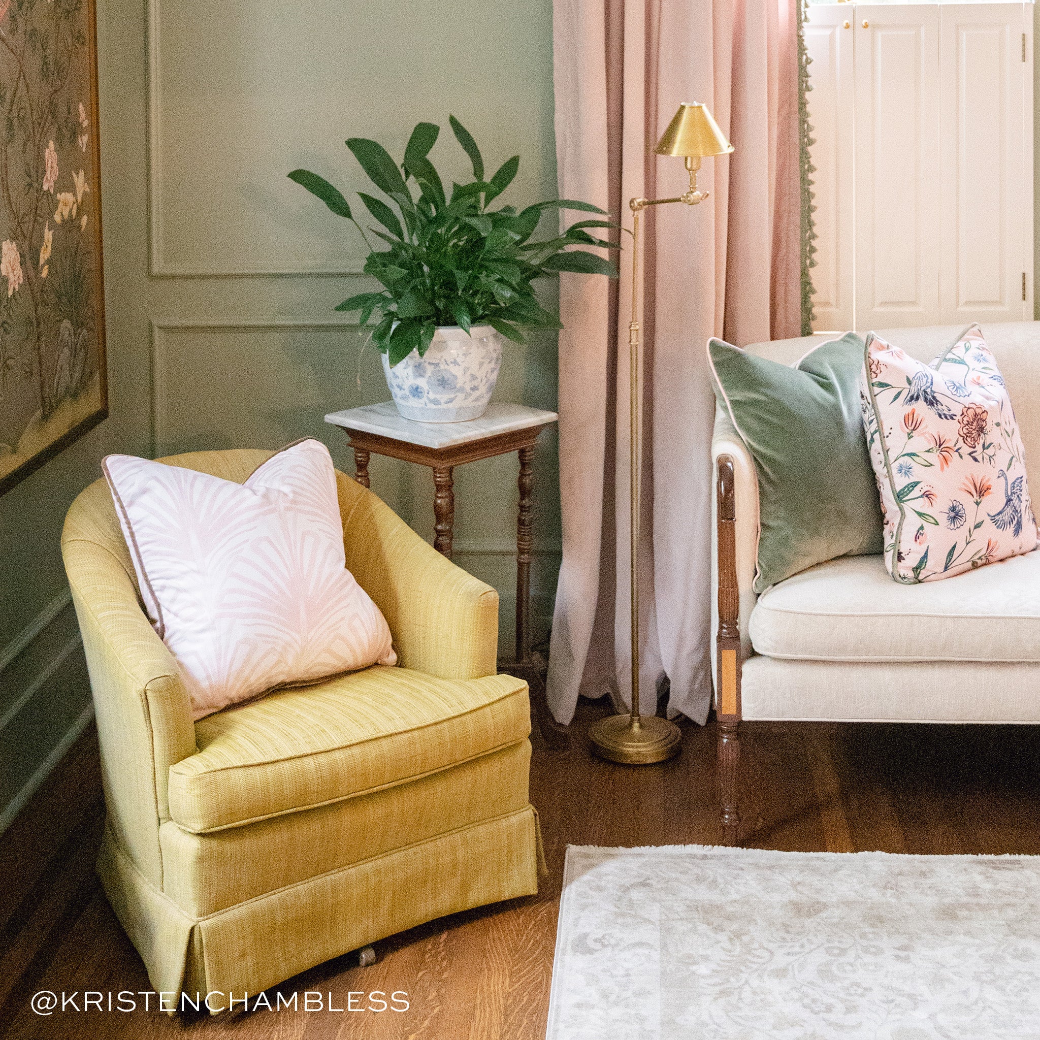 Living room corner styled with Rose Pink Art Deco Printed Pillow on yellow sofa couch and Fern Green Velvet and Pink Chinoiserie Printed Pillow on cream couch next to Pink Curtains with Sage Pom Poms by marble wooden table with plants in white and blue vase on top. Photo taken by Kristen Chambless