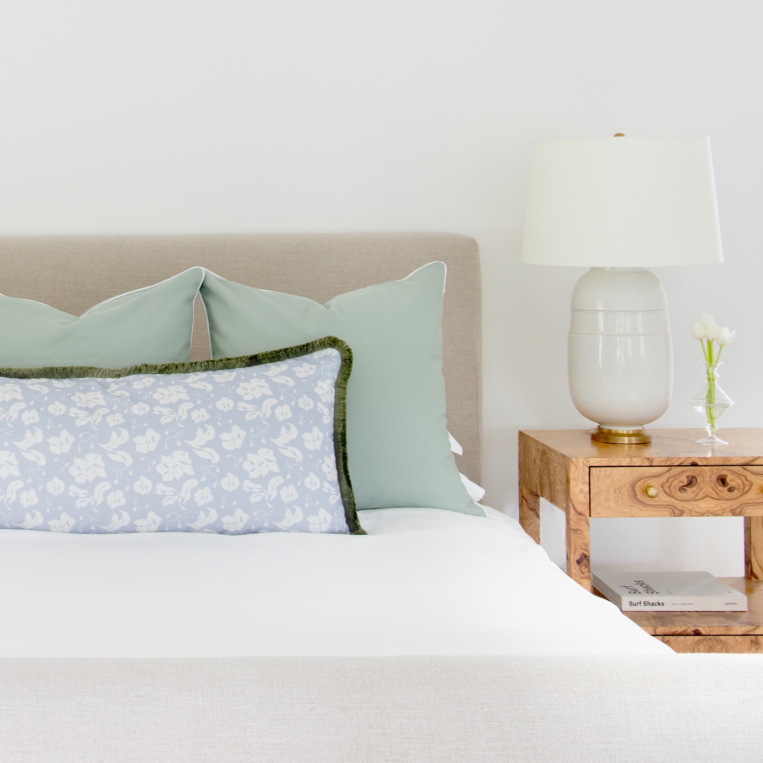 White bed close-up styled with sage green pillows and one Cornflower Blue Floral Lumbar with sage fringe next to light brown nightstand with white lamp and clear vase with white flowers on top