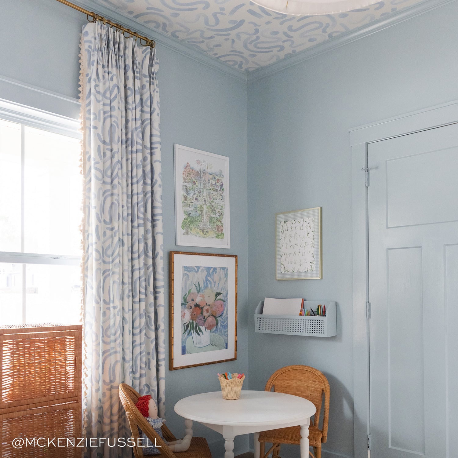 sky blue abstract curtains hung on a metal rod in front of an illuminated window in a room with light blue walls and sky blue patterned wallpaper on the ceiling