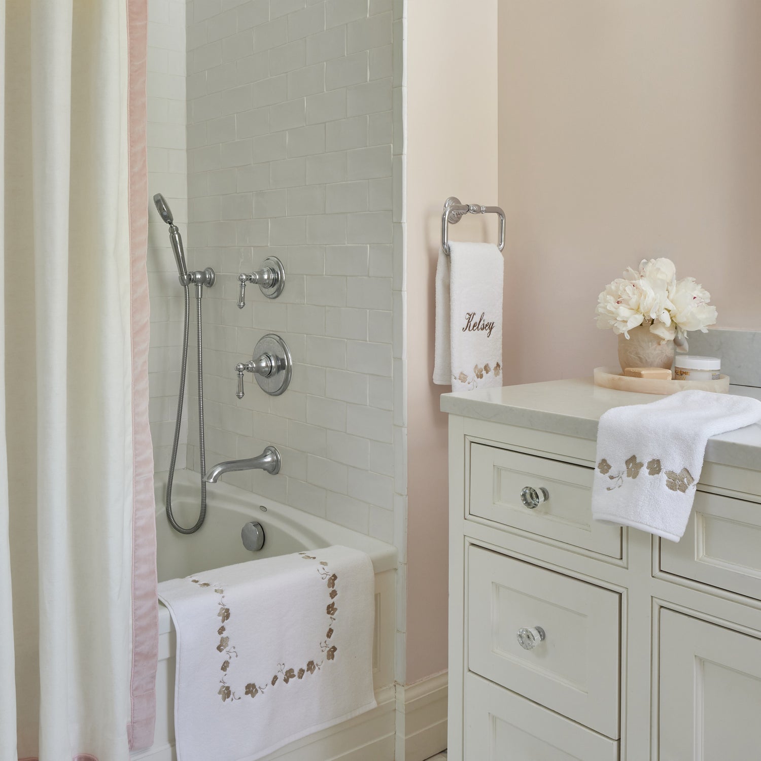 white towel and bath mat with light brown embroidered floral trim in a pink bathroom with a white shower curtain hung with a light pink velvet band and white tiles in the tub