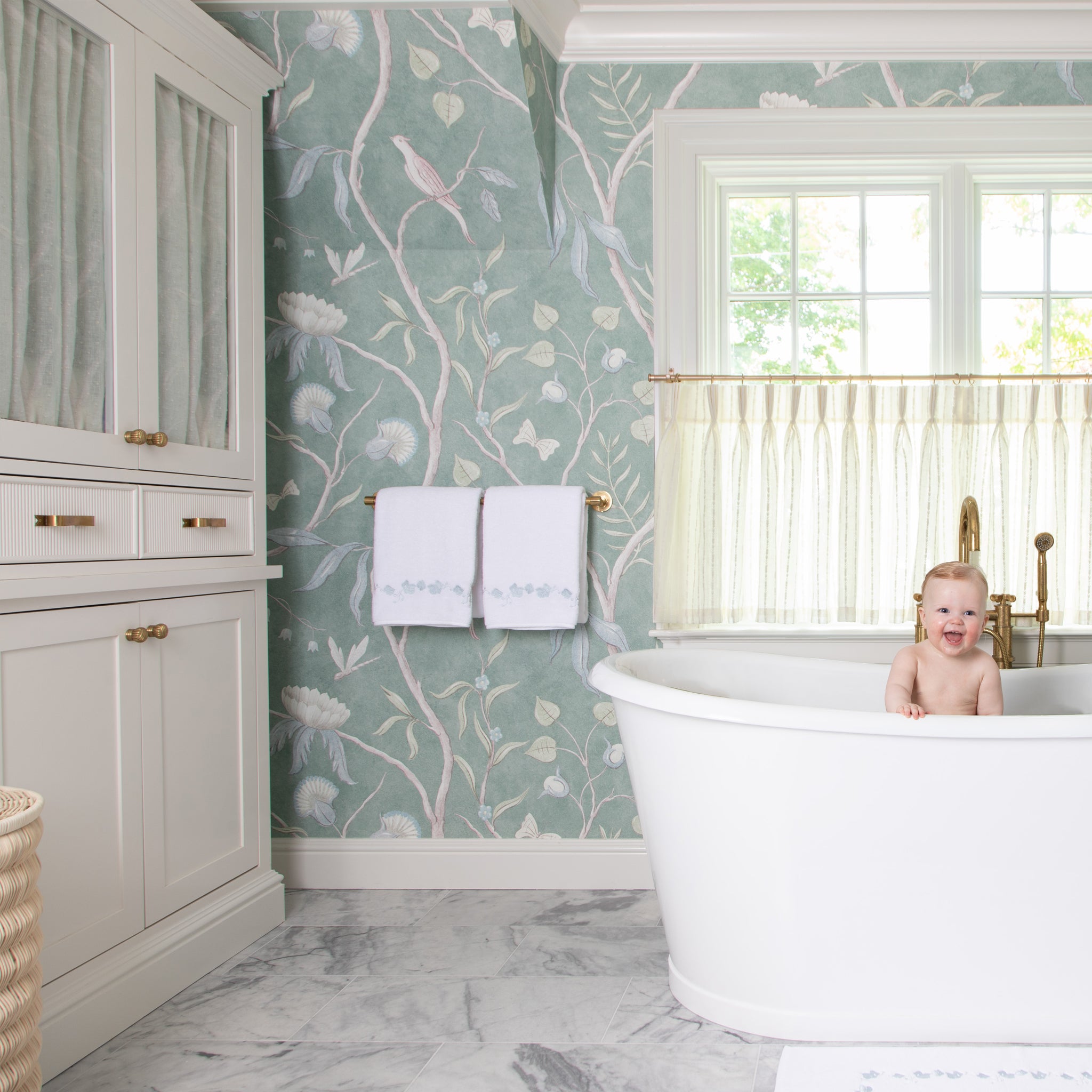 blue botanical wallpaper on the walls in a bathroom with a white bath tub with a baby in the tub and an illuminated window behind the tub with neutral cream cafe curtains hung in it and white towels with blue botanical embroidery hung on a gold metal rod next to the tub