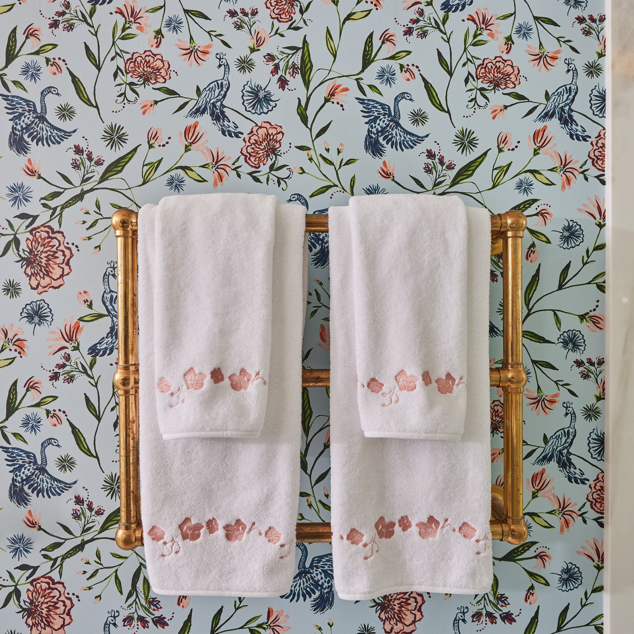 Powder blue chinoiserie wallpaper on a wall in a bathroom with a gold metal towel rack with white towels on it with embroidered coral botanical stripe