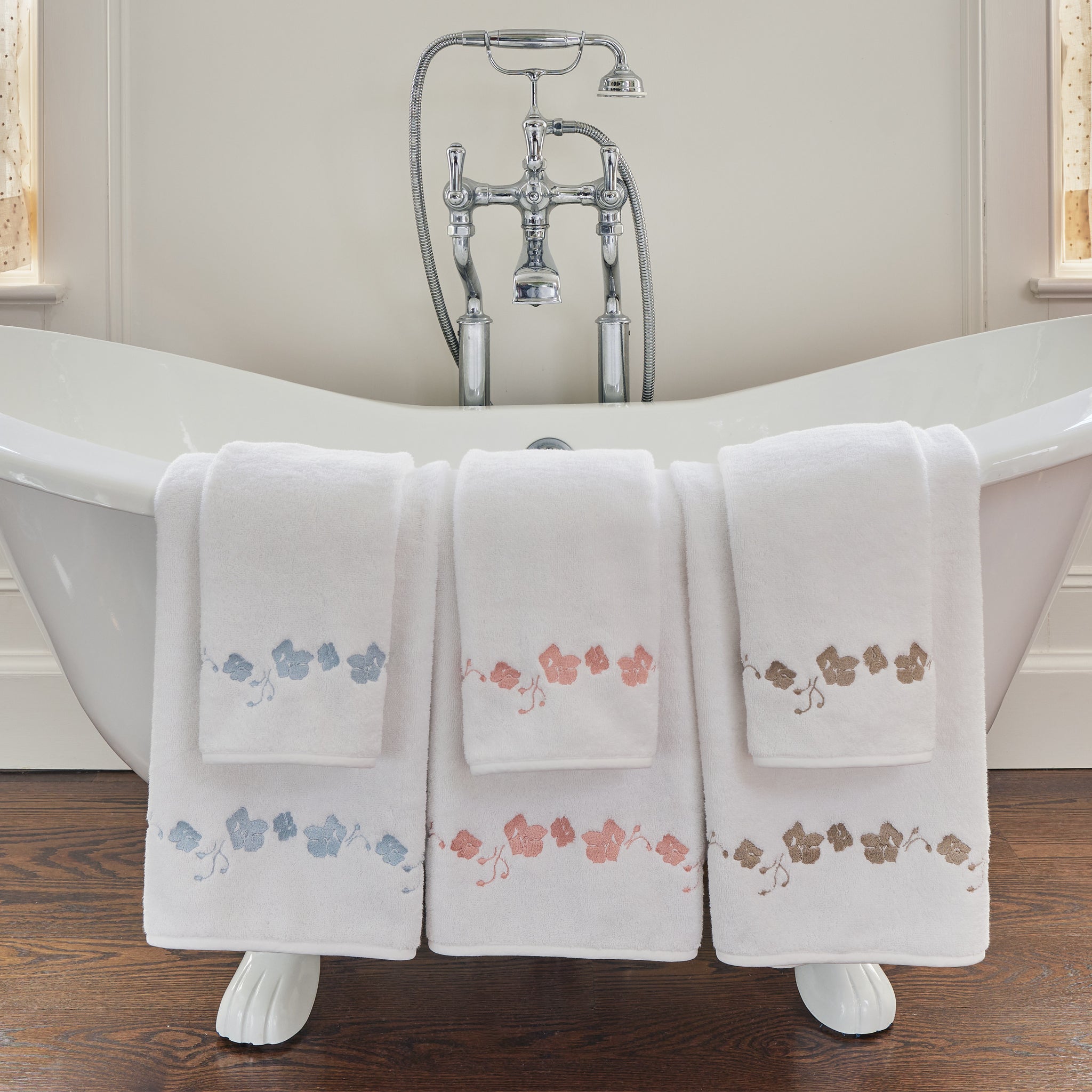 a white bathtub with a set of blue embroidered botanical stripe towels, coral embroidered botanical stripe towels, and sand embroidered botanical stripe pattern hung over the side of the bath tub