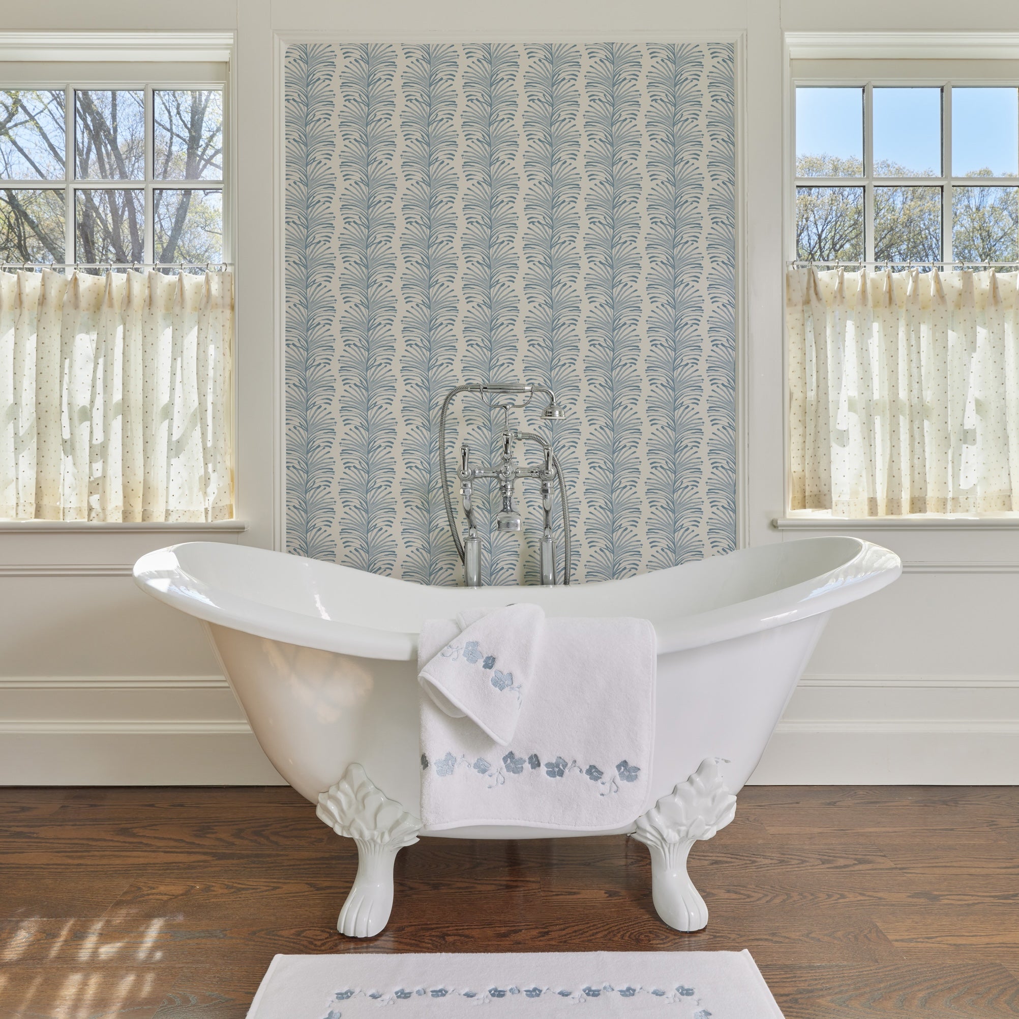 bathroom with Sky Blue Botanical Stripe wallpaper on the walls, cream cafe curtains on the two windows, a white bathtub in the middle of the image with white towels with and embroidered blue botanical stripe on them and a white embroidered bath mat with blue botanical pattern