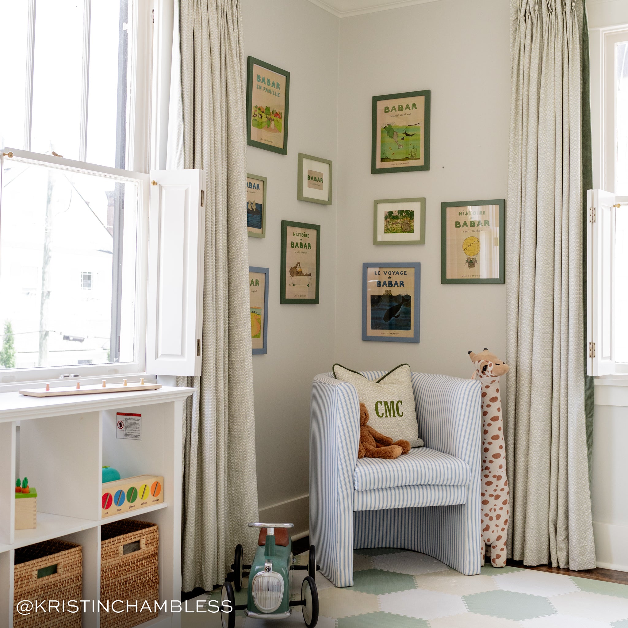 close up of a corner of a play room with white walls and moss green geometric curtains hung in front of illuminated windows with a moss green geometric pillow sitting on a blue and white striped chair in the corner with framed pictures hung on the wall above the chair and toys and stuffed animals on the ground