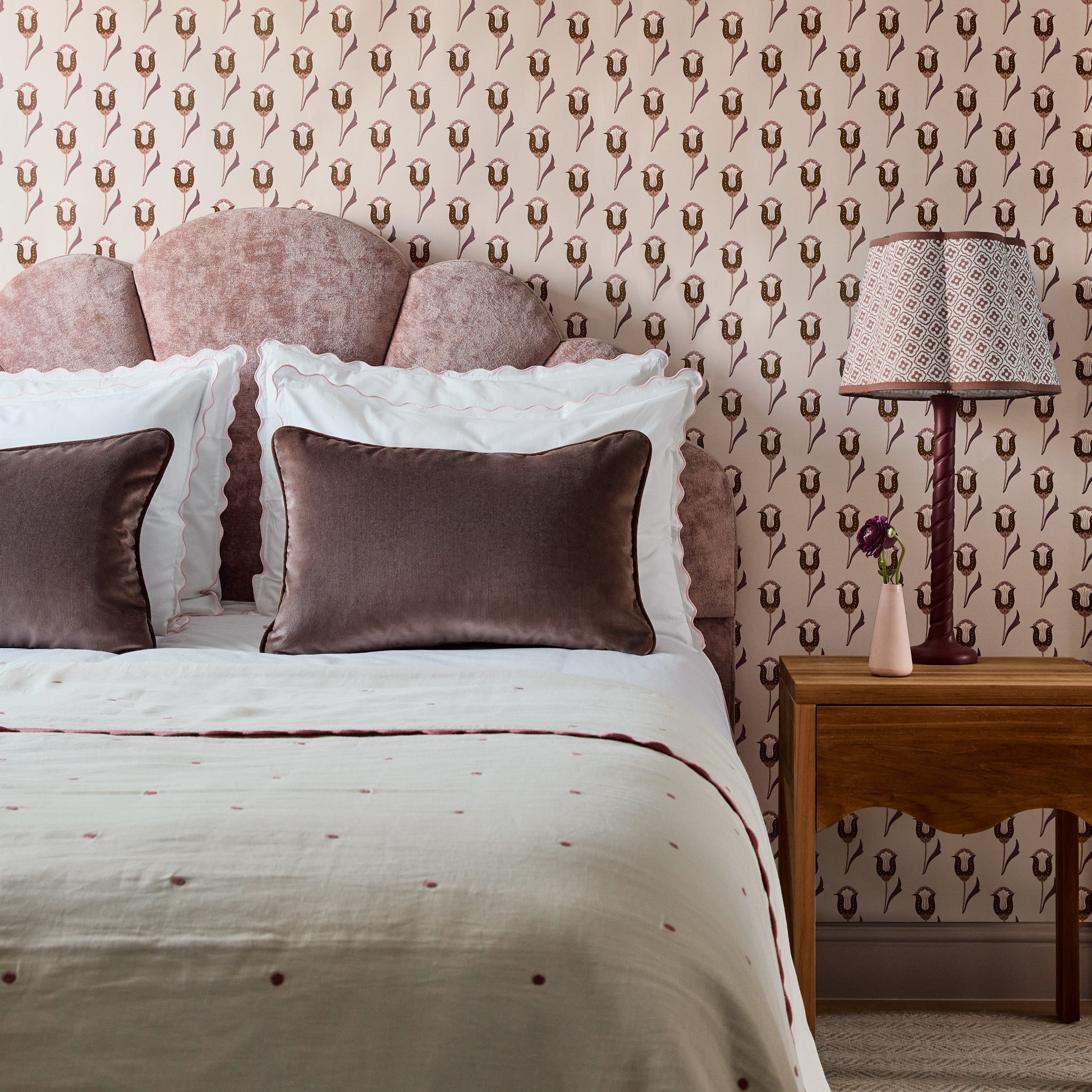 abstract floral neutral background wallpaper with pink and maroon flowers on it and a bed in front of the wall with white polka dot bedding and brown velvet pillows on the bed and a brown wooden night stand next to the bed with a lamp on it