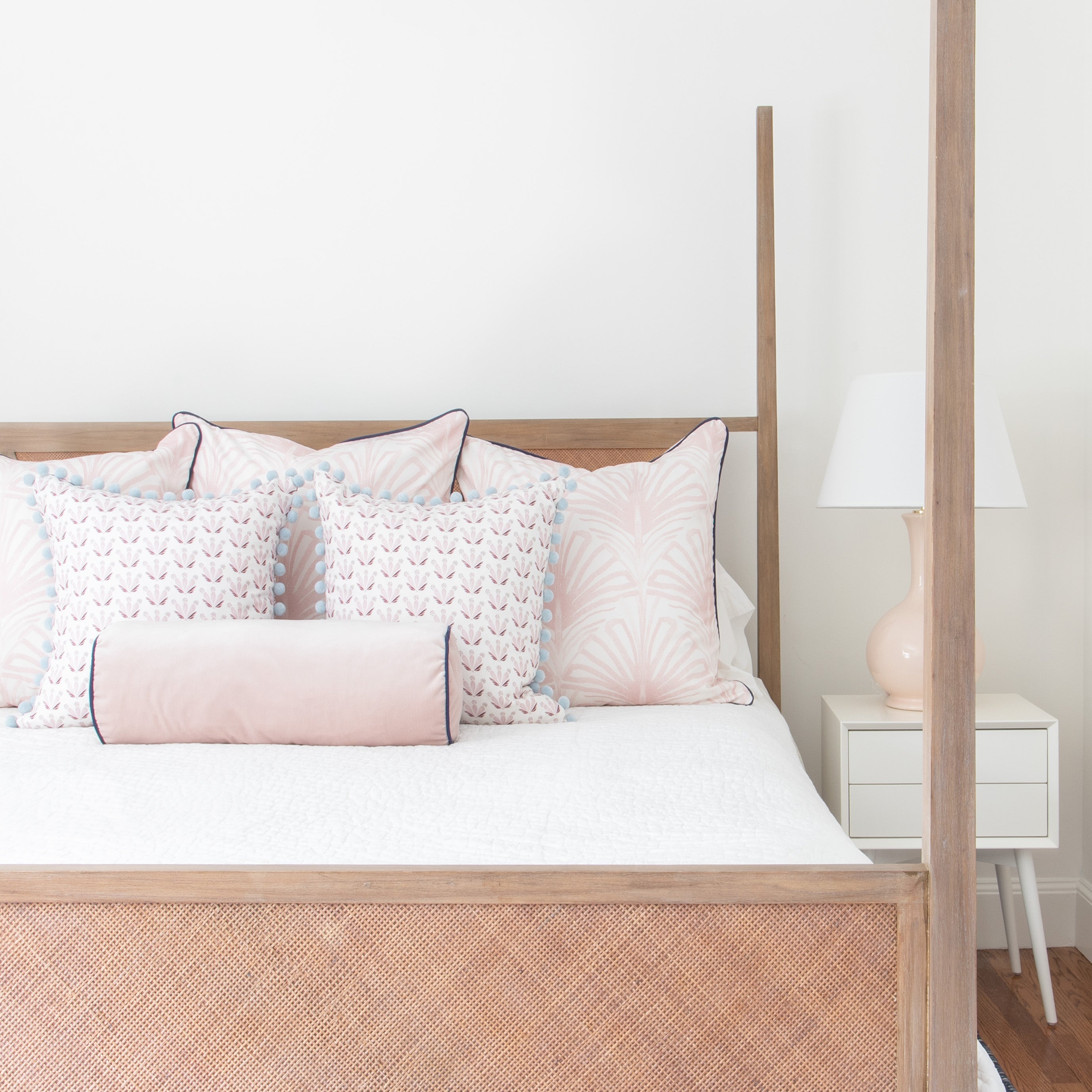 Wooden Bed styled with three Rose Pink Art Deco Printed pillows, two Pink & Burgundy Drop Repeat Floral Printed pillows, and one Pink velvet Lumbar next to white nightstand with white and pink lamp on top