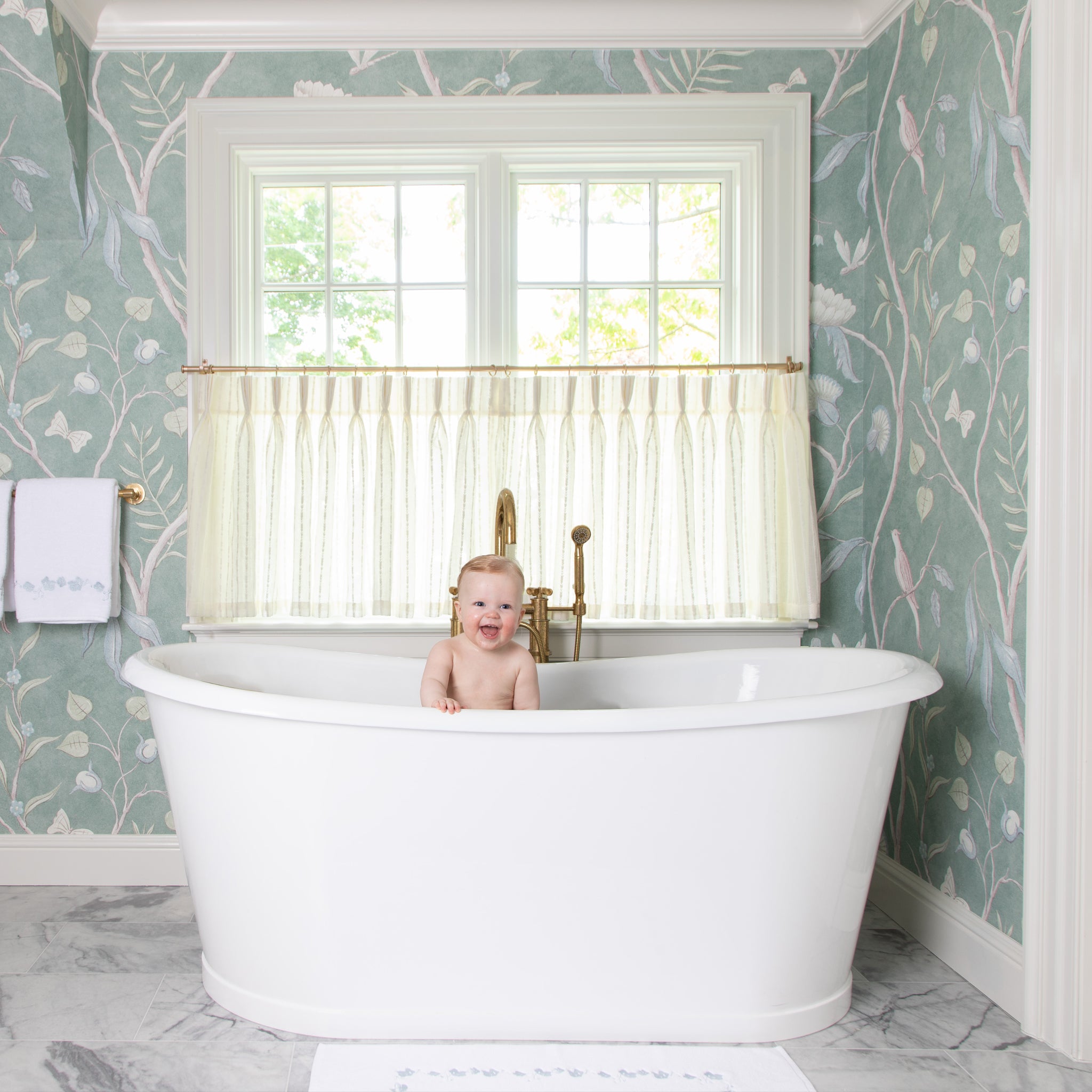 sheer white cafe curtain with embroidered white stripes hung in an illuminated window in a bathroom with a white bath tub in front of the window with botanical wallpaper on the walls
