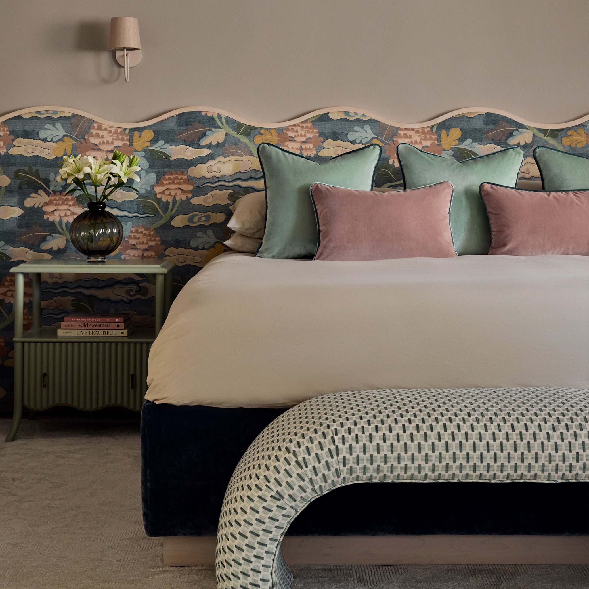 A cozy bedroom featuring a scalloped, floral-patterned upholstered headboard, pastel throw pillows in pink and green, a beige duvet, and a curved patterned bench. A side table holds a vase with white flowers and books, complemented by a wall sconce above.