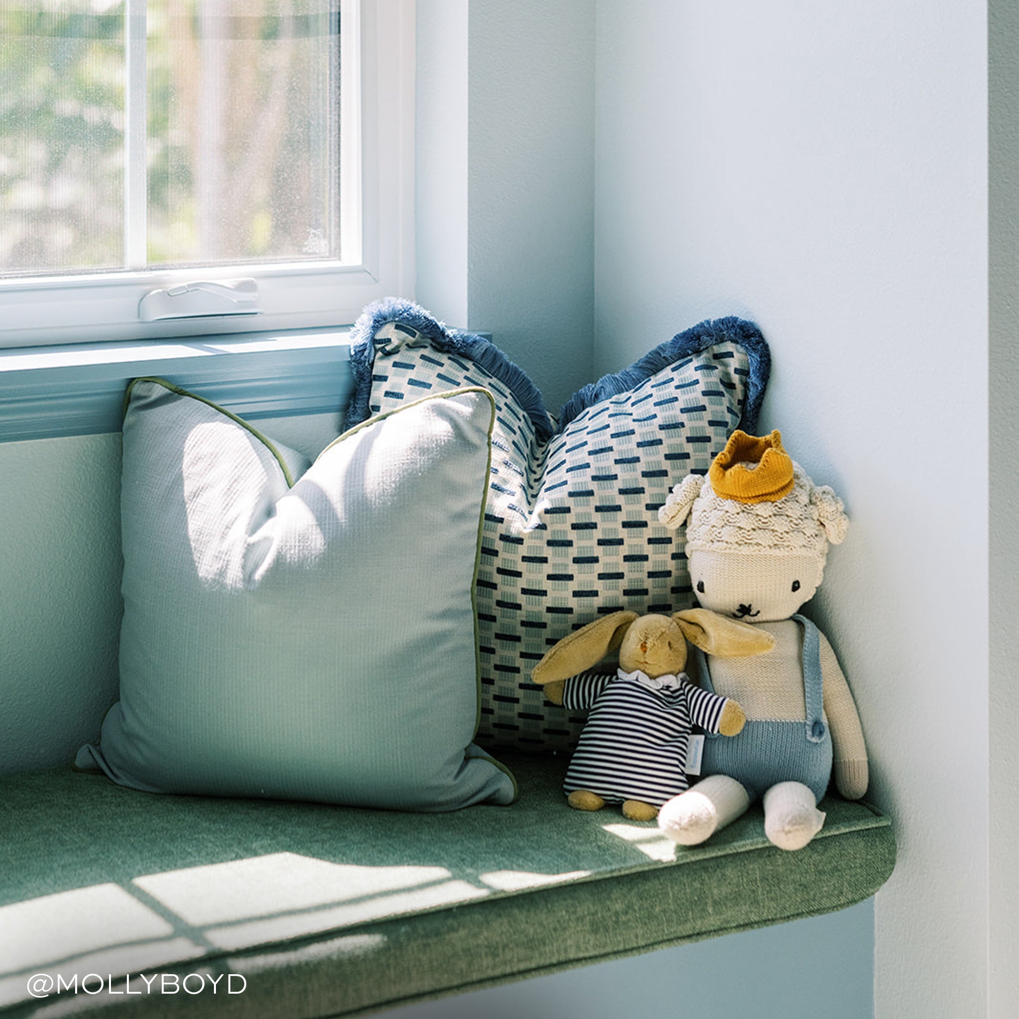 blue grey pillow with green piping and a woven blue chenille jacquard pillow with blue fringe sitting on a green window bench with stuffed animals in front of the pillows