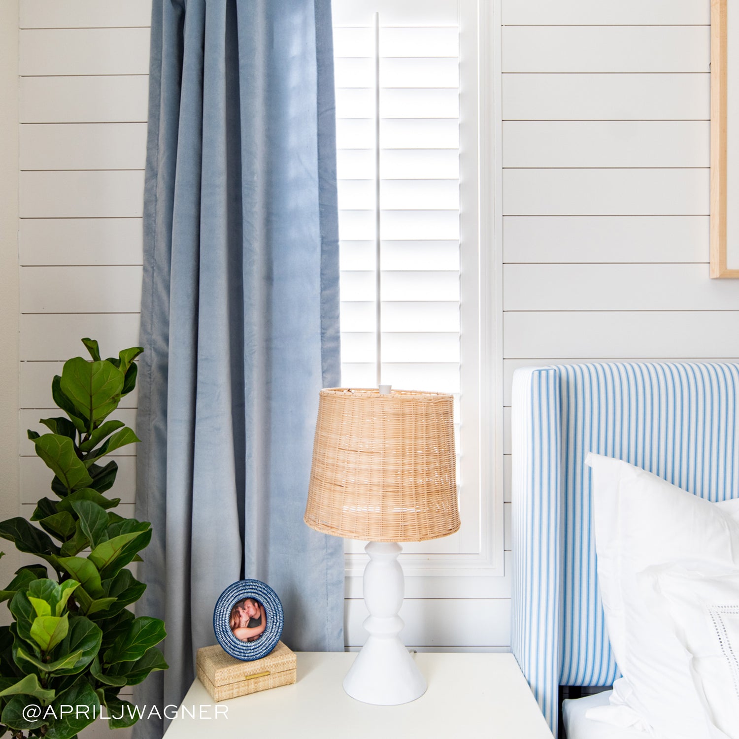 Close-up of Nightstand with white lamp on top next to window styled with sky velvet curtains. Photo taken by April J Wagner