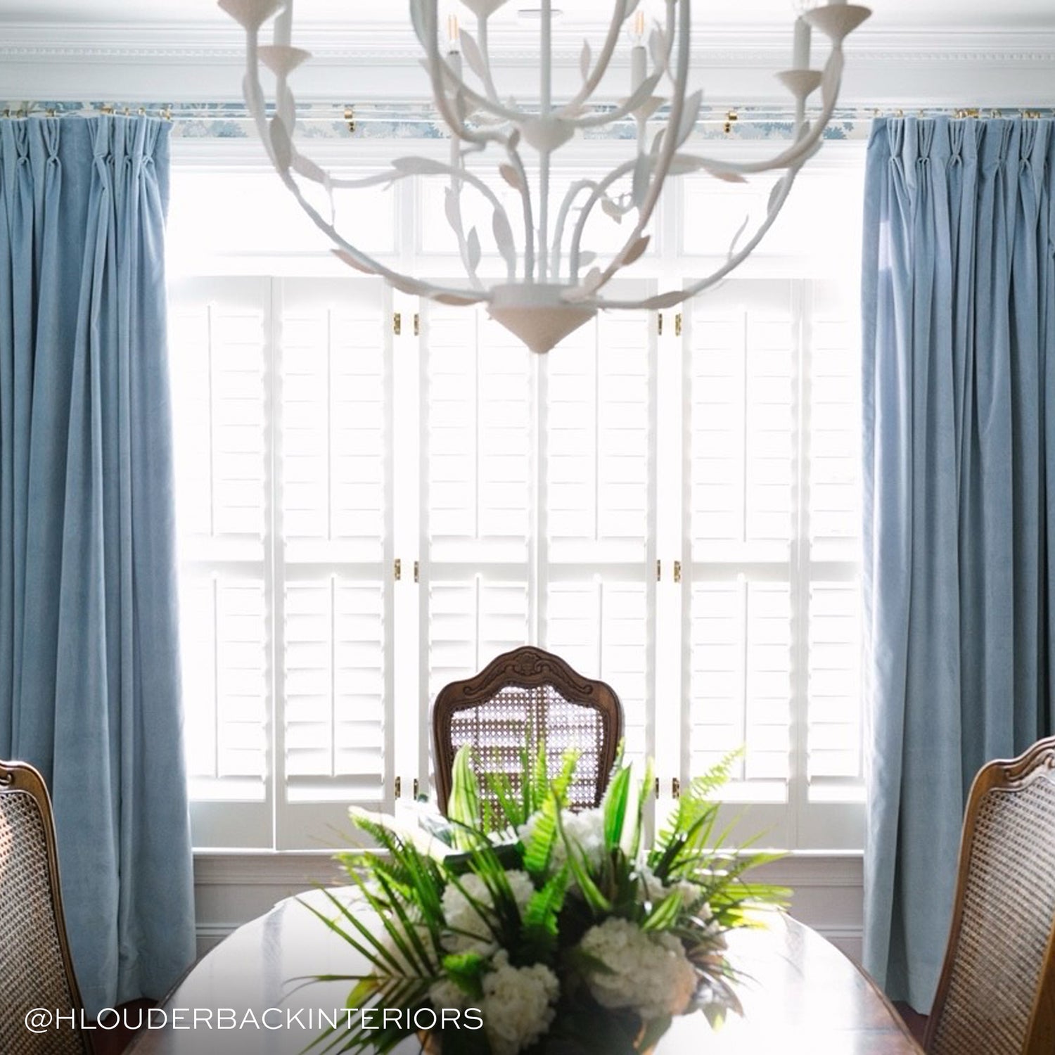 photo of a dining room with a white chandelier hanging, blue velvet curtains hung in front of an illuminated window and a brown wooden table with a floral arrangement on it