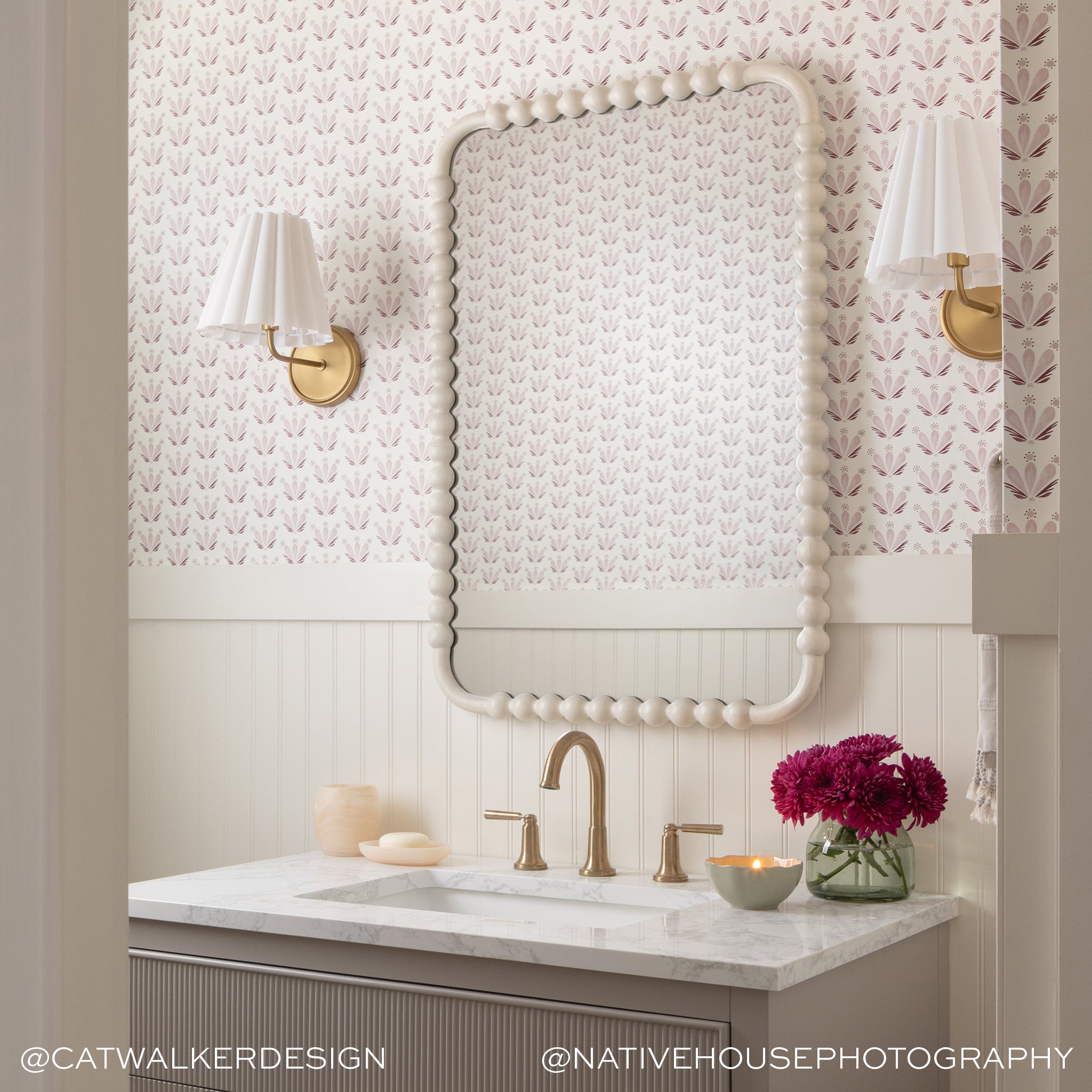Bathroom vanity with Serena Pink floral-patterned wallpaper, custom mirror, brass fixtures, and fresh flowers in a vase