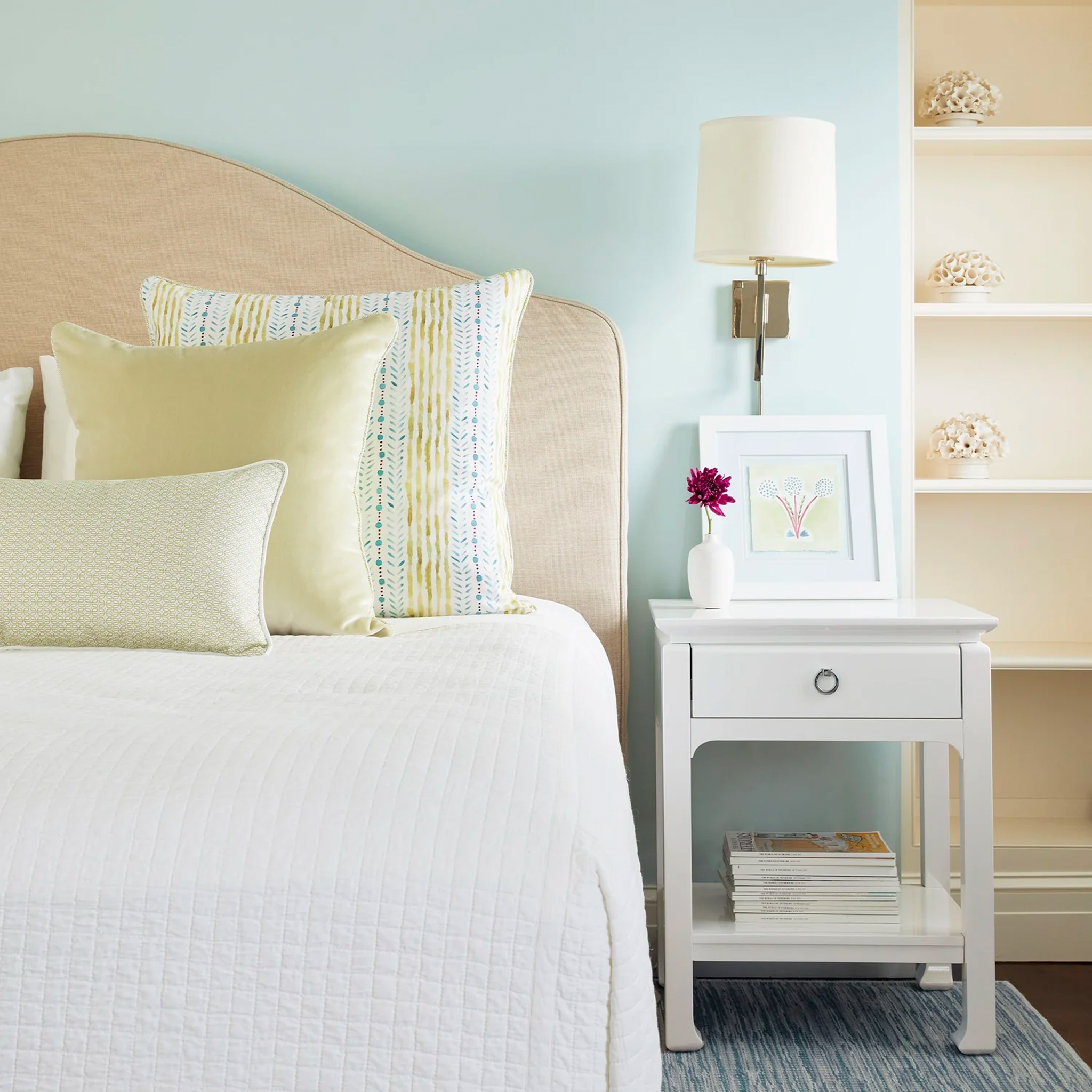 White bed close-up styled with Blue & Green Striped Printed Pillow, Light Green Velvet Pillow, and Moss Green Geometric Printed Pillow next to white nightstand with white frame and pink flower in white vase on top