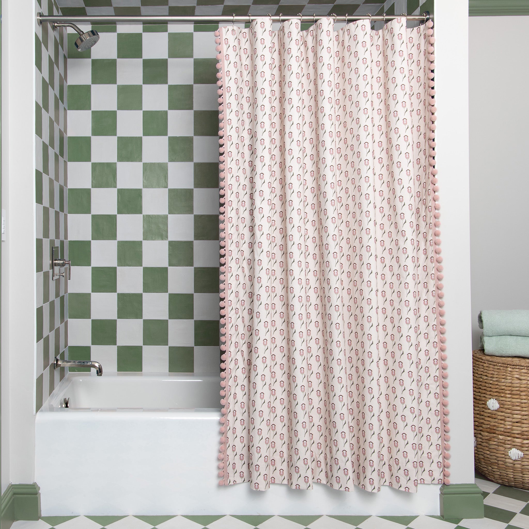 abstract pink and green floral patterned shower curtain with pink pom poms hanging in a bathroom shower with green and white tiles