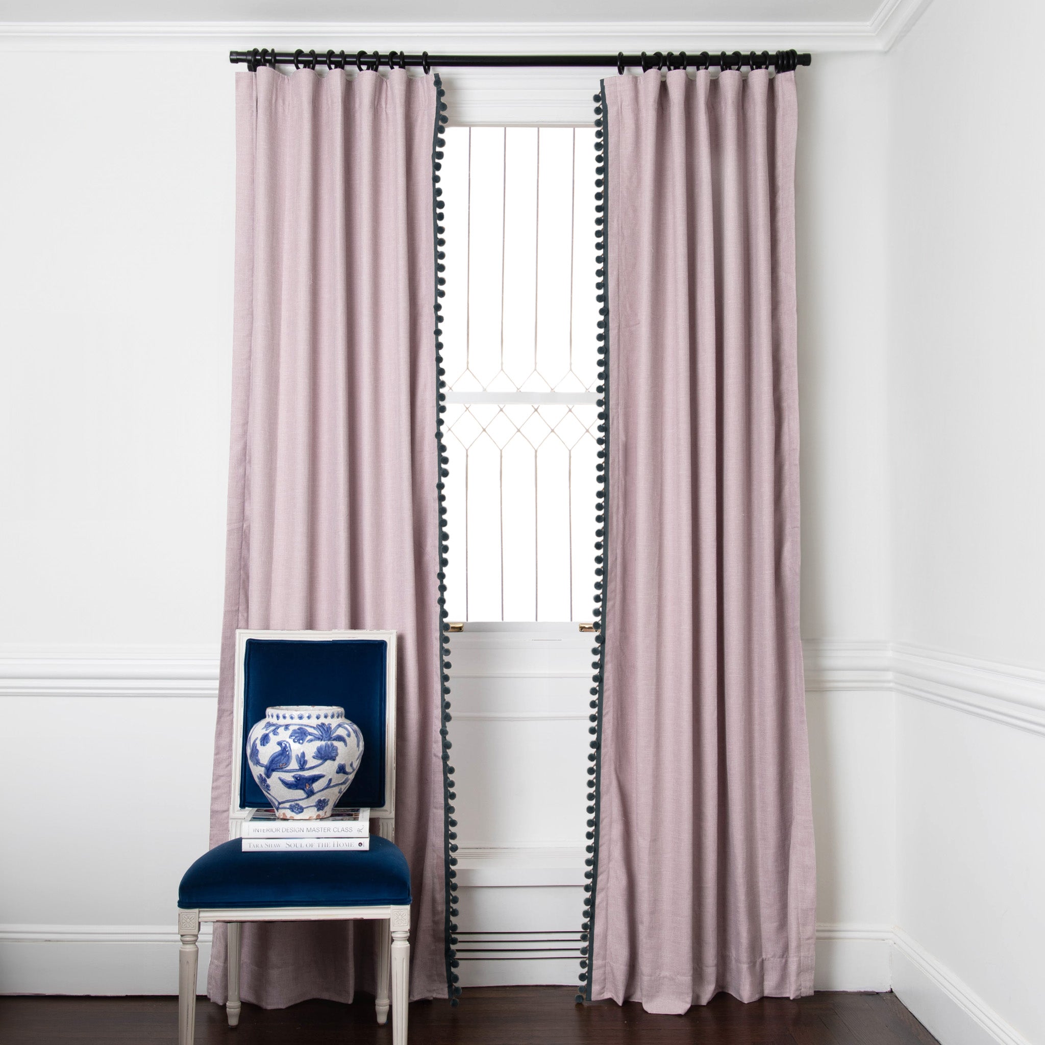 pink curtains on a metal rod in front of an illuminated window with a navy chair in front stacked with books
