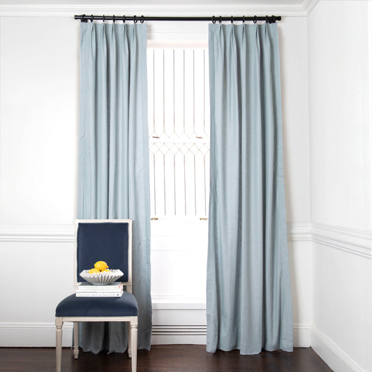 grey blue curtains on a metal rod in front of an illuminated window with a navy blue chair in front stacked with books