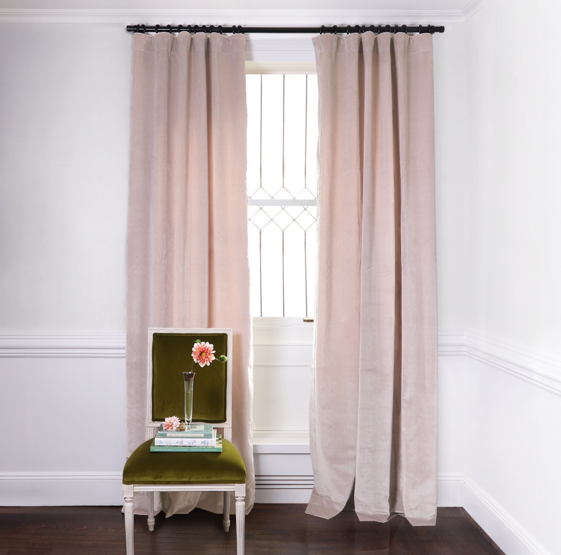 Pink Velvet Curtains on black rod in front of an illuminated window with Moss Green Velvet chair with flowers in clear vase on top of two stacked books