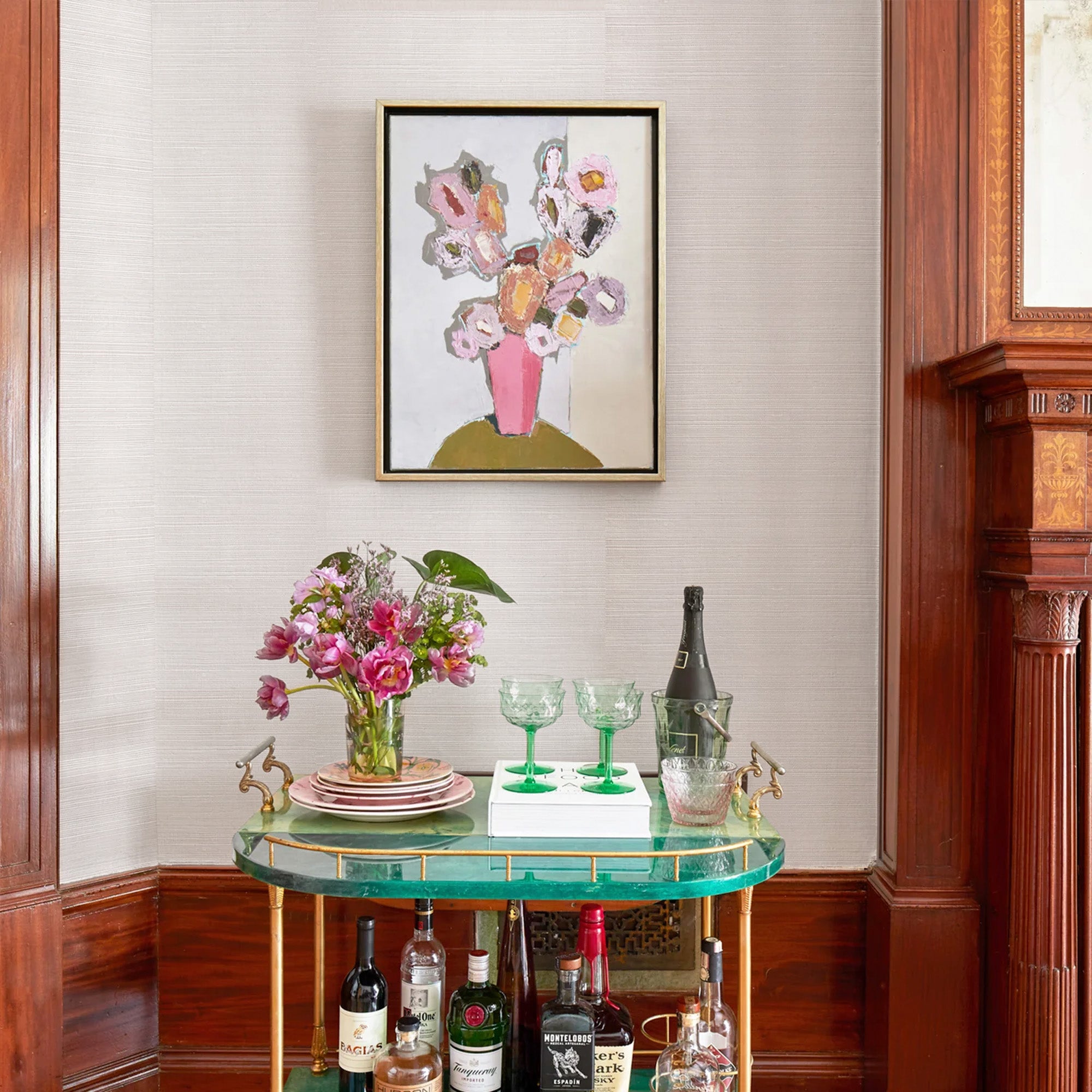 wall with Pink Grasscloth Wallpaper and a painting of flowers hung on the wall and a green bar cart with flowers and green glasses on it