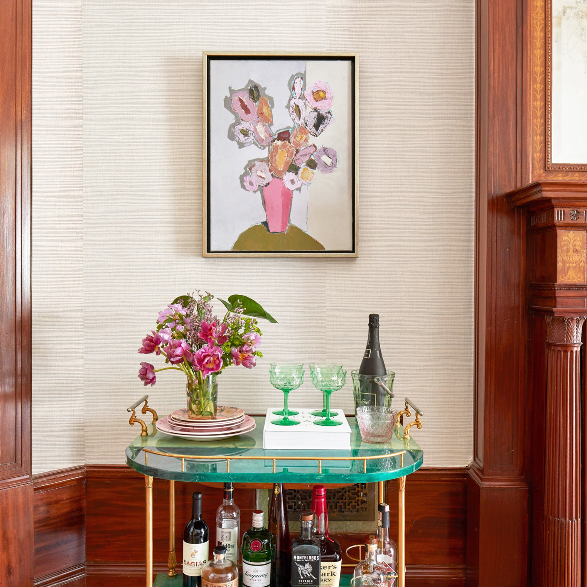 wall with ivory grasscloth wallpaper and a painting of flowers hung on the wall and a green bar cart with flowers and green glasses on it