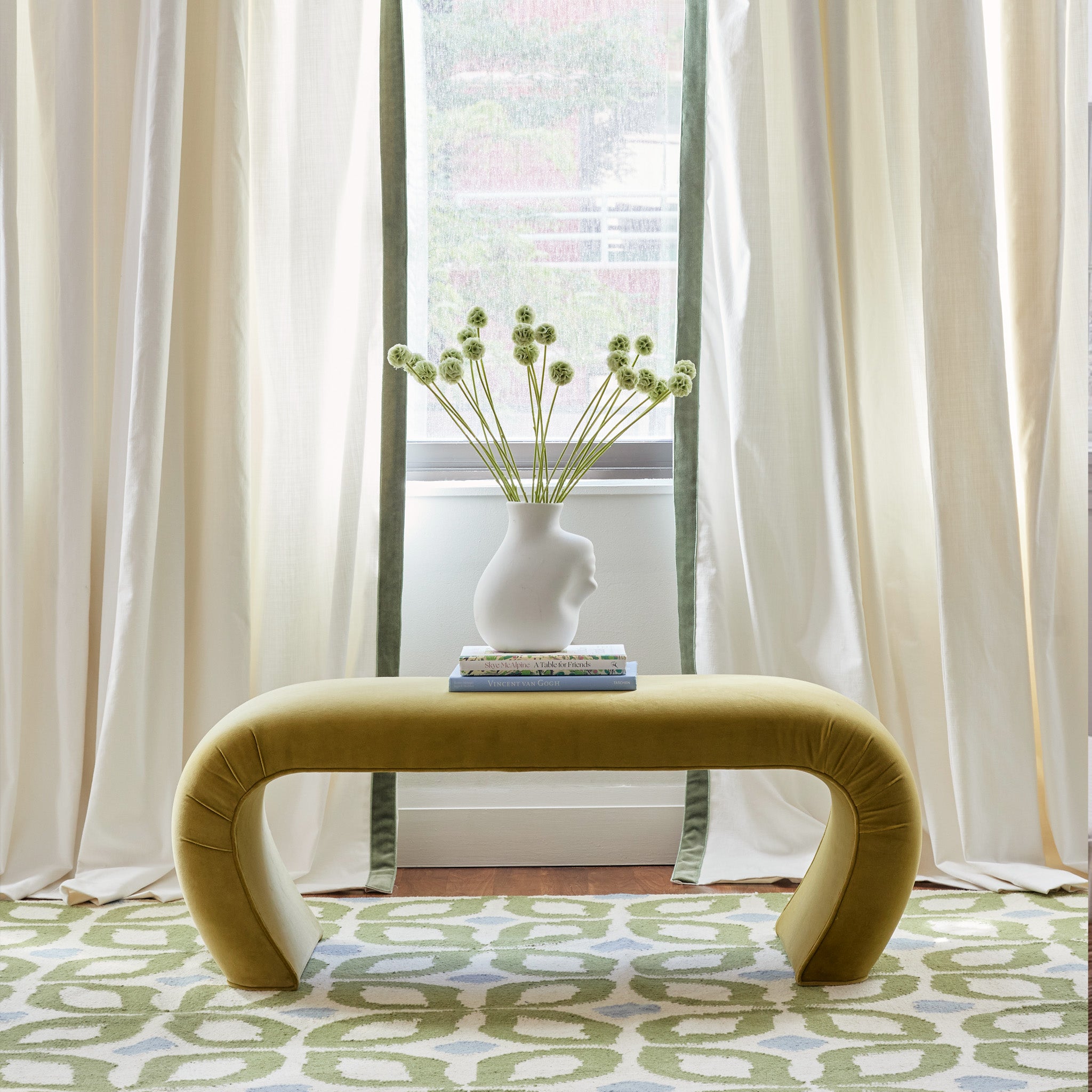 white curtains with a sage green velvet band hung in front of an illuminated window with a geometric green and blue rug and a dark yellow table with books and a vase on it