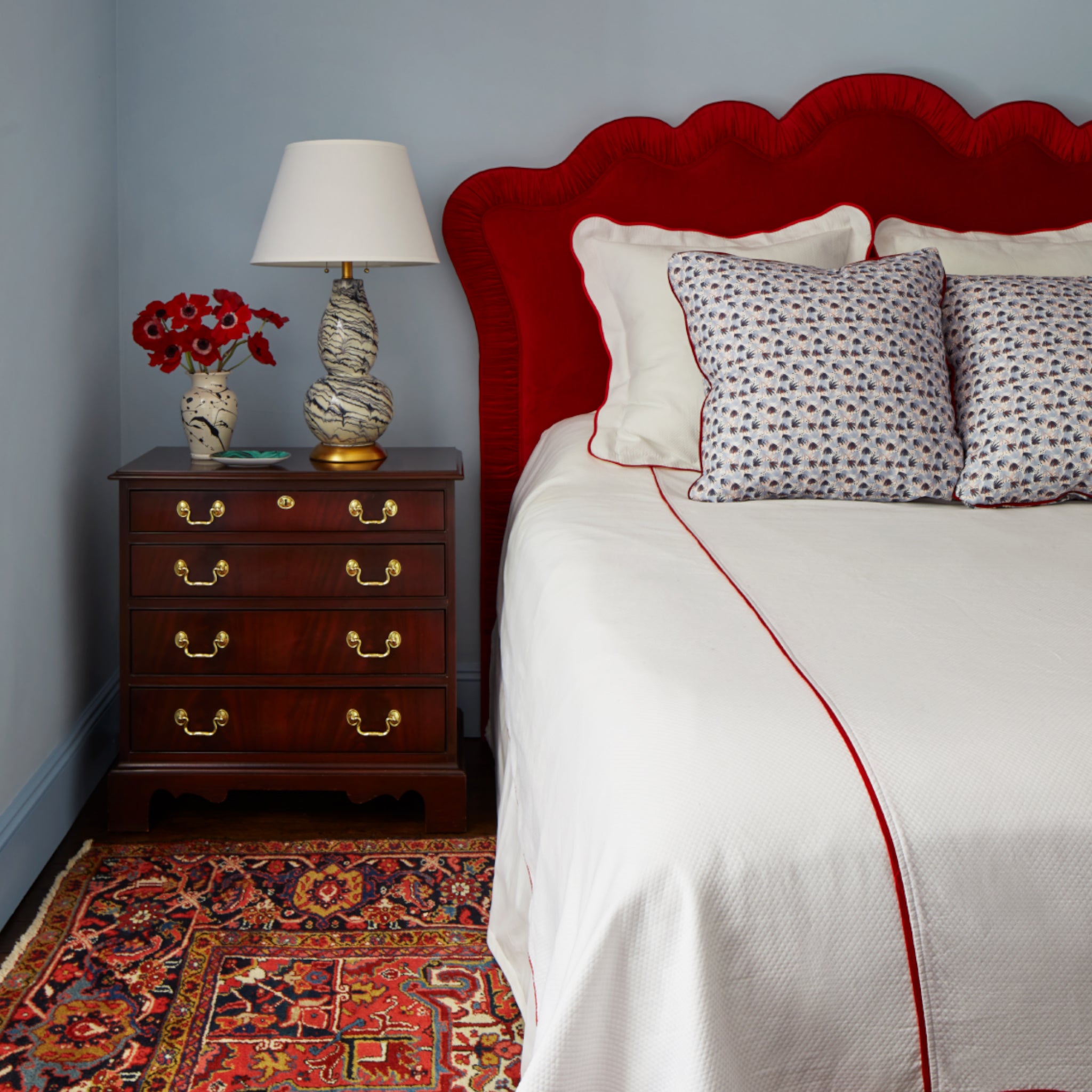 Bedroom styled with Red and Blue Printed Pillows on White bed with red headboard next to a wooden nightstand with white lamp and red flowers in vase on top