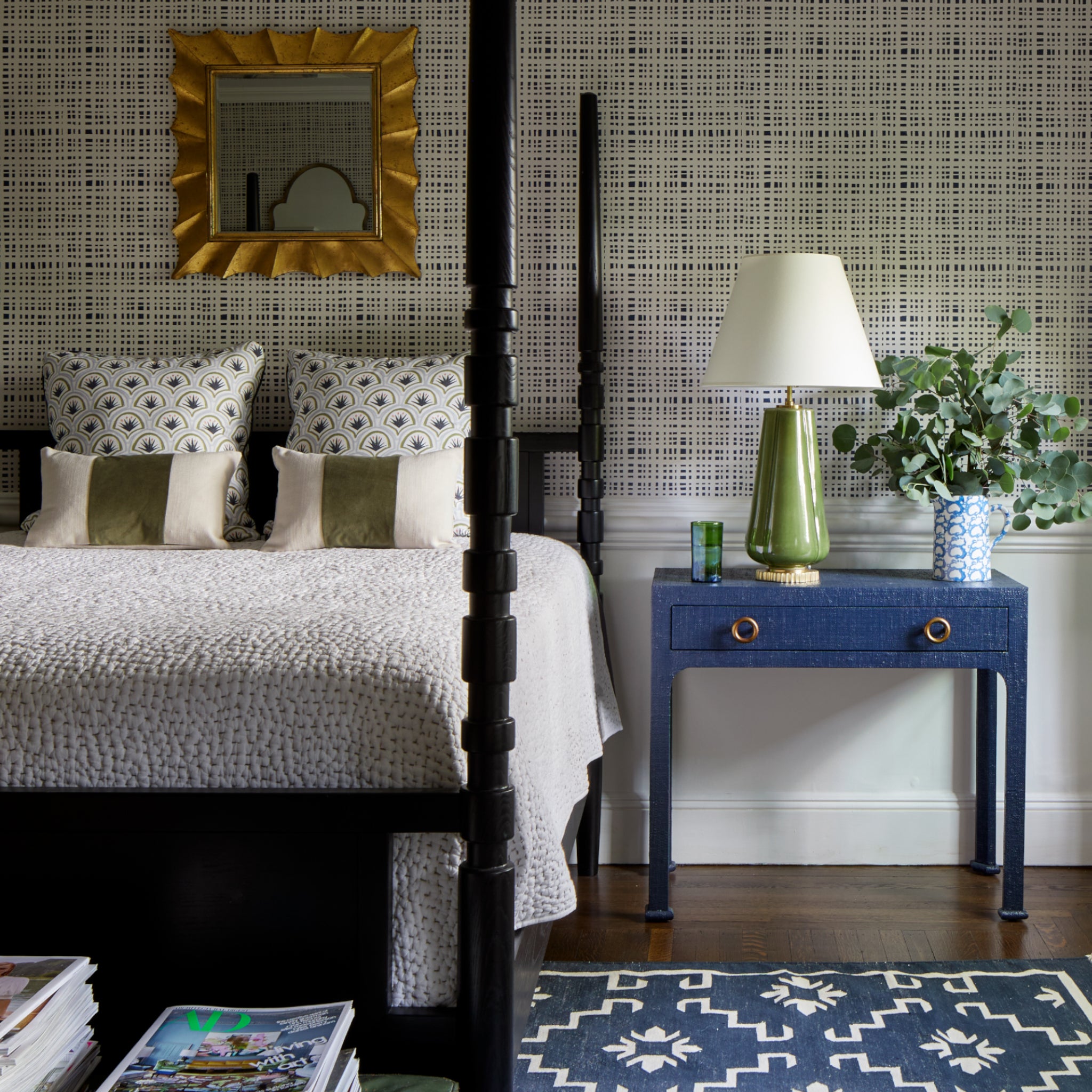 Bedroom styled with two Art Deco Palm Pattern printed pillows and two white and green lumbars on white bed next to blue table with lamp and plants in vase on top over a navy gingham printed wallpaper