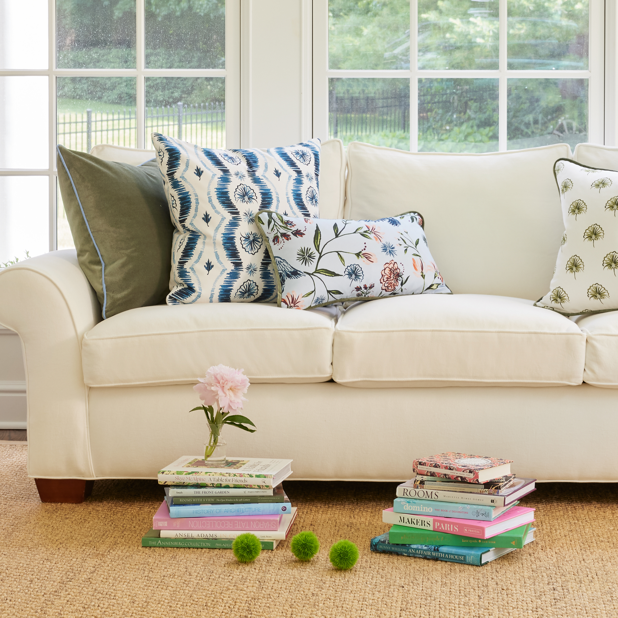 White couch on living room with fern green pillow, blue ikat pillow, daphne powder pillow, and green floral pillow by two stacks of books on the floor with one stack having a pink flower on top with white framed window in the back