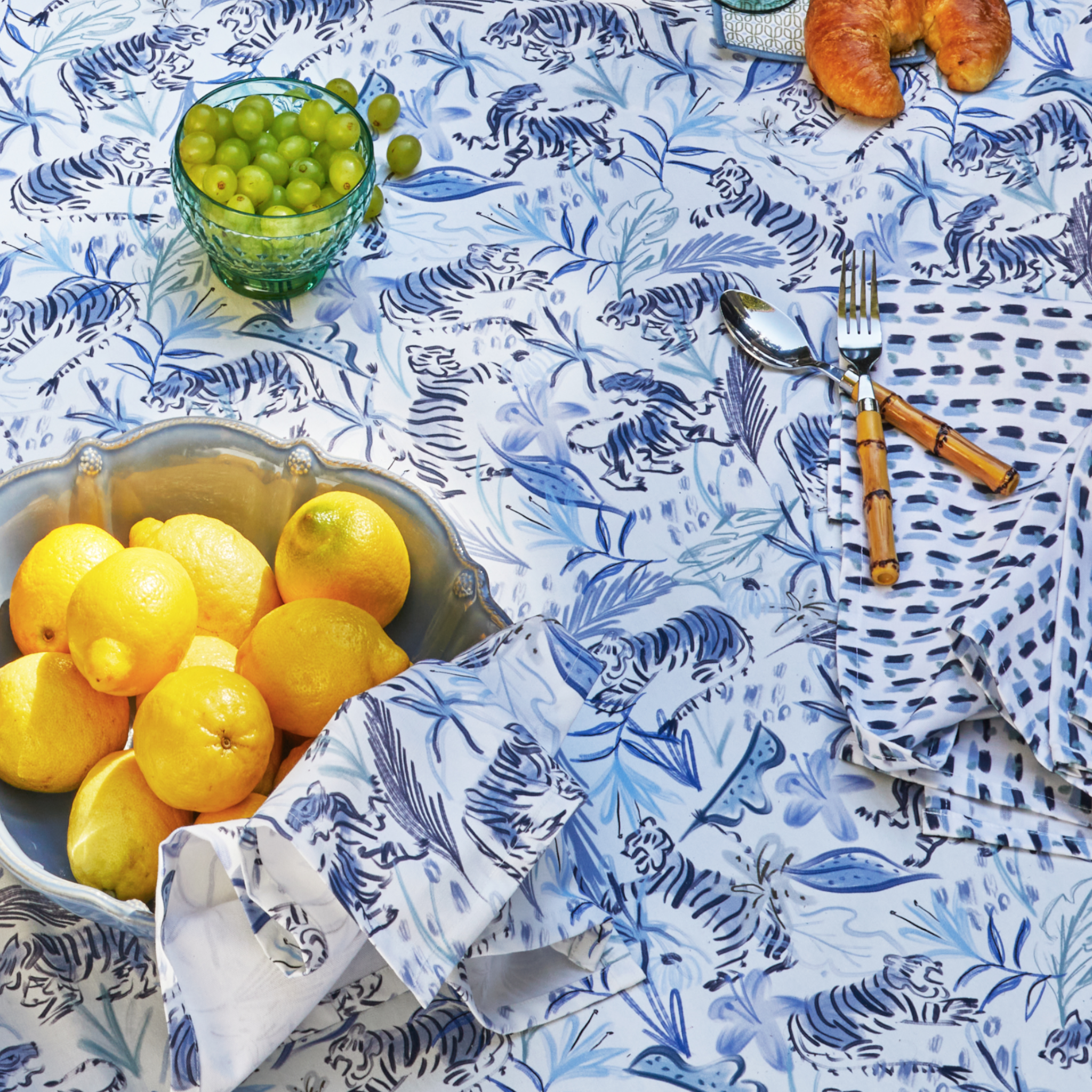 Table Close-Up with Blue With Intricate Tiger Design Printed Tablecloth and lemon bowl with Blue With Intricate Tiger Design Printed Napkin on top by glass of grapes