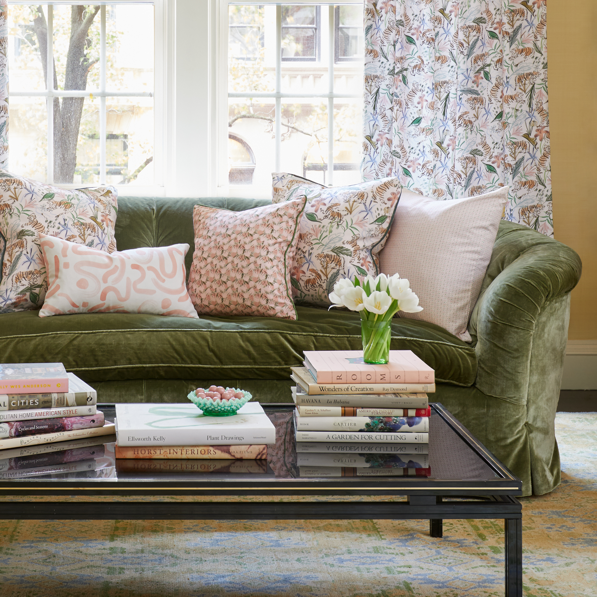Living room styled with a Pink Geometric printed pillow, Pink Chinoiserie printed pillow, Pink Floral printed pillow, and Pink Graphic printed lumbar on Fern Green Velvet couch by coffee table with stacked books and white tulips in vase in front of an illuminated window with Cream Chinoiserie printed curtains