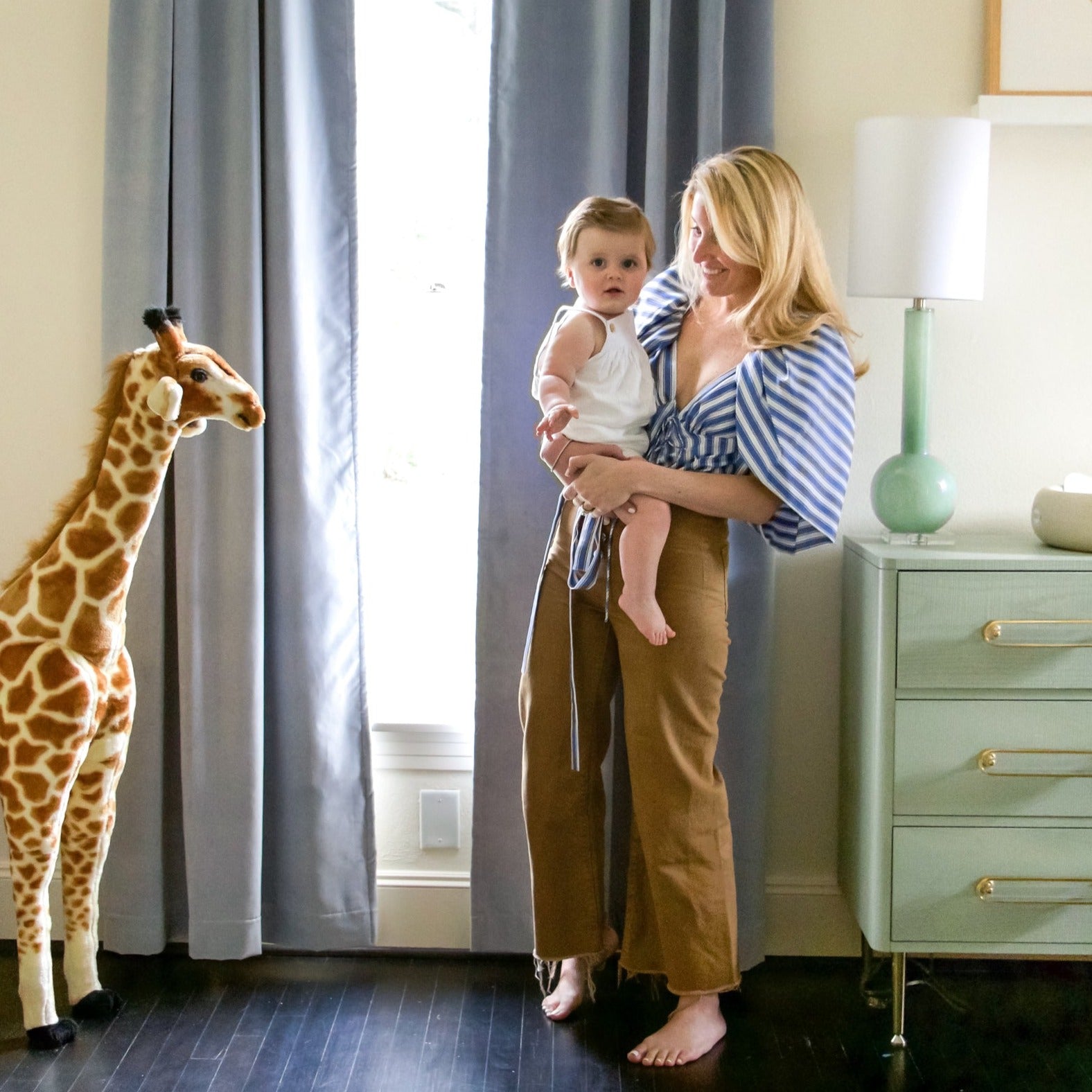 Nursery room window styled with sky velvet curtains and blue green dresser next to blonde joyful mother carrying blonde baby
