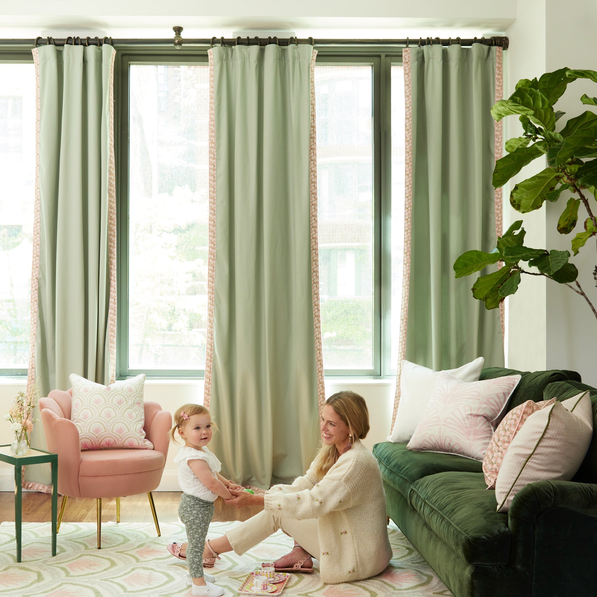 Living room styled with Light Green Curtains with Pink Floral Band by Rose Pink Art Deco Printed Pillow, Pink Floral Printed Pillow, and Pink Geometric Printed Pillow next to mother and baby child holding hands on Pink Art Deco Palm Printed Rug with Pink Art Deco Palm Printed Pillow on pink velvet sofa chair by small green table with pink flowers in clear vase