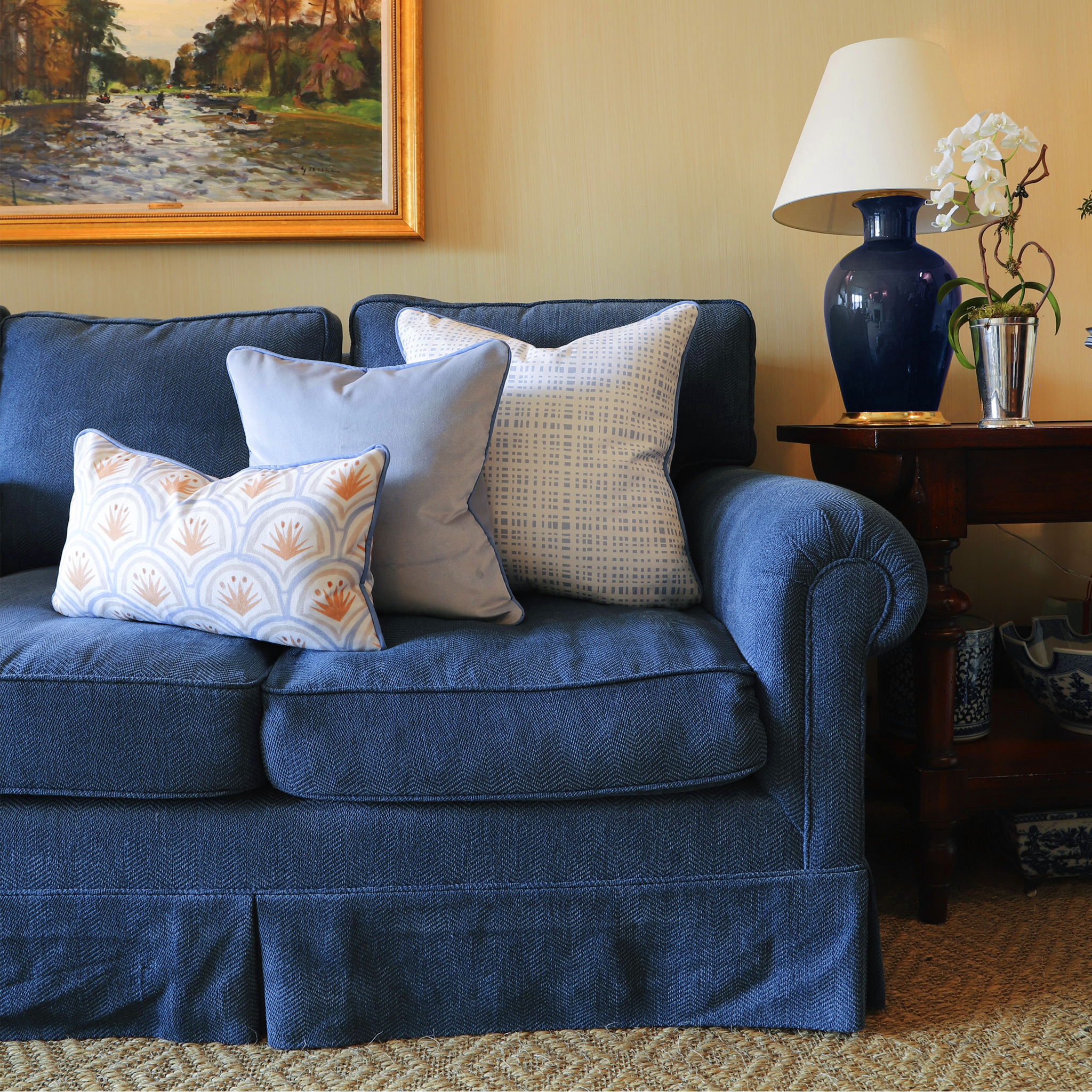 Living room close-up of blue couch with Art Deco Palm Pattern Printed Lumbar, Sky Blue Velvet Pillow, and Sky Blue Gingham Printed Pillow next to wooden table with blue and white lamp next to white flowers in silver vase