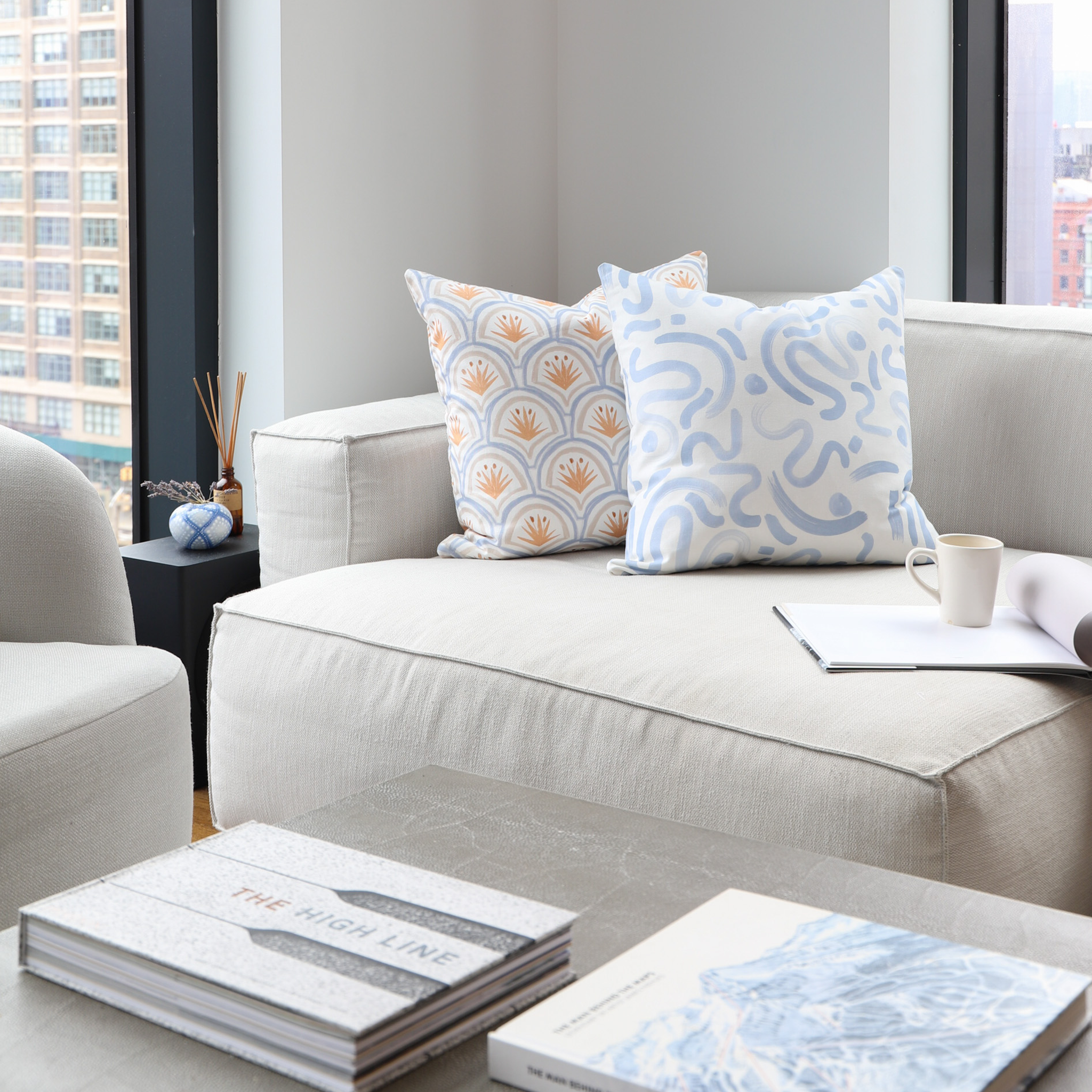 Oat Linen Couch Close-Up styled with an Art Deco Palm Pattern Printed Pillow and a Sky Blue Printed Pillow by window with books on coffee table in front 