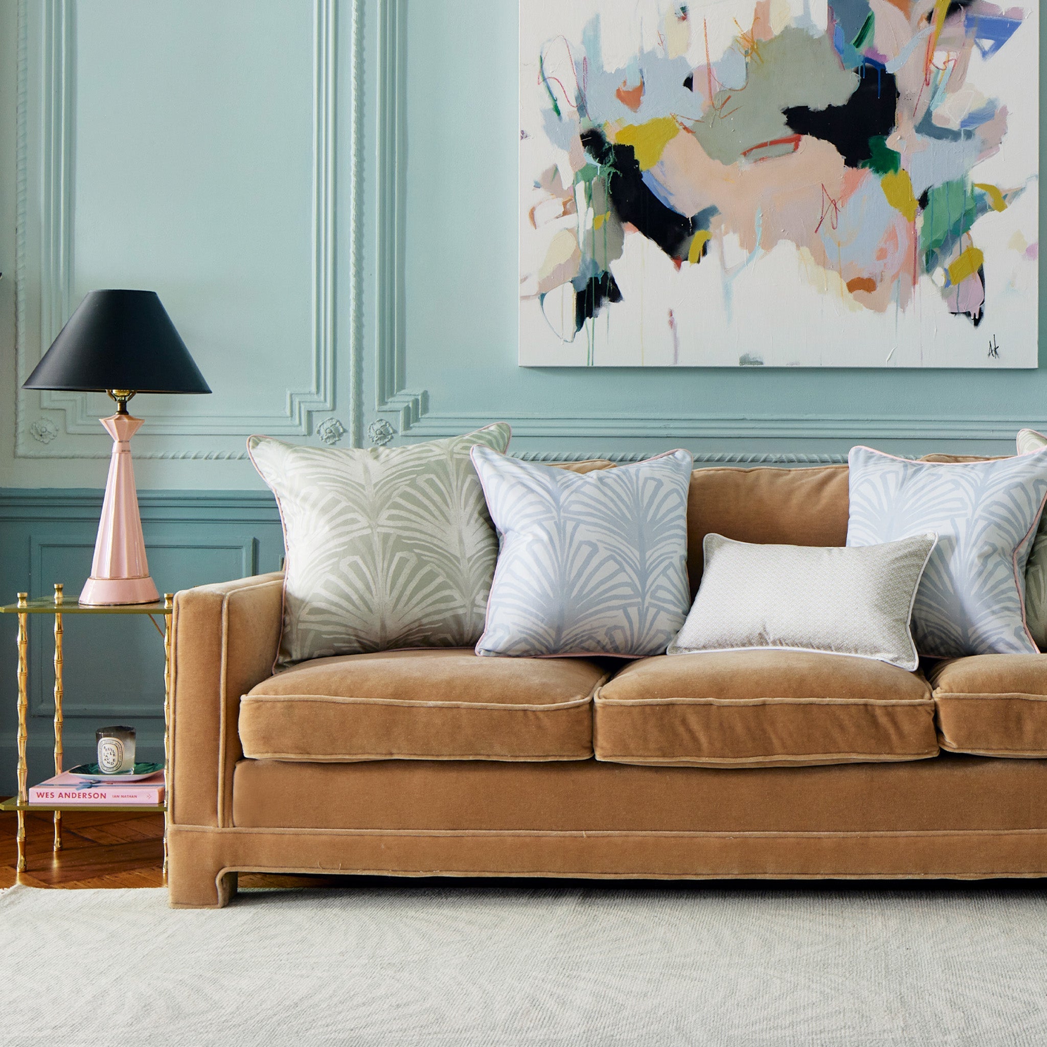 Close-up of light brown velvet couch styled with one Sage Green Printed Pillow, two Sky Blue Palm printed pillows, and one Moss Green Geometric Printed Lumbar next to a glass table with pink and black on top