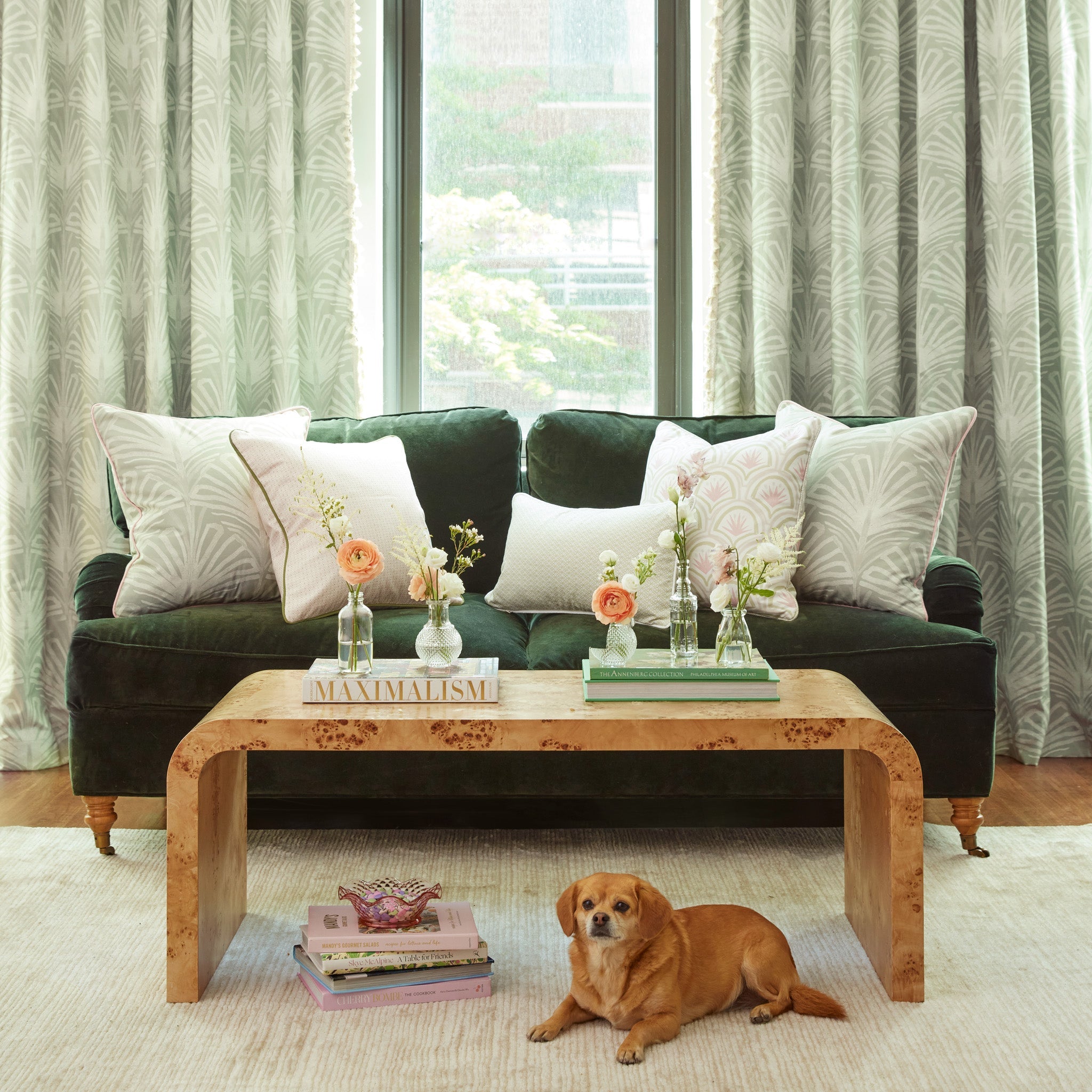 Living room styled with Sage Green Palm Printed Curtains and two Sage Green Palm Printed Pillows, one Pink & Green Palm Printed Pillow, one Pink Geometric Printed Pillow, and one Moss Green Geometric Lumbar on fern green velvet couch next to light brown coffee table with books and flowers in vases on top and books stacked on floor next to brown dog over rug