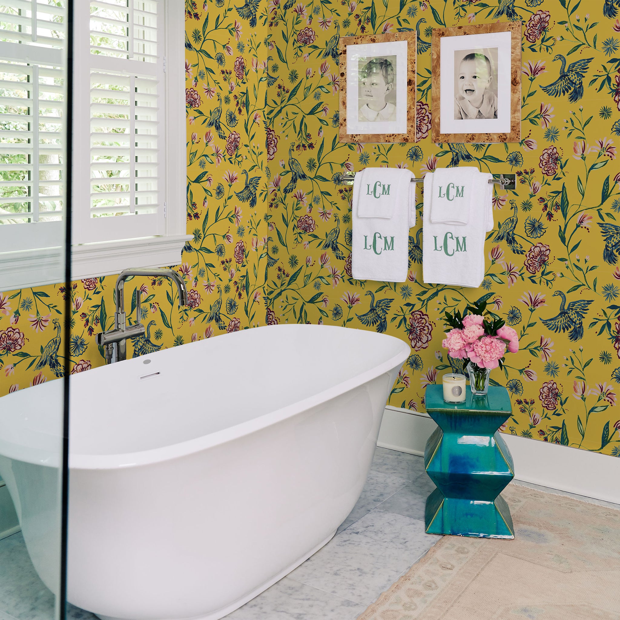 Bathroom styled with Yellow Canary printed wallpaper with white bath tub and two photo frames hanging from wall on top of the hanging monogrammed towels