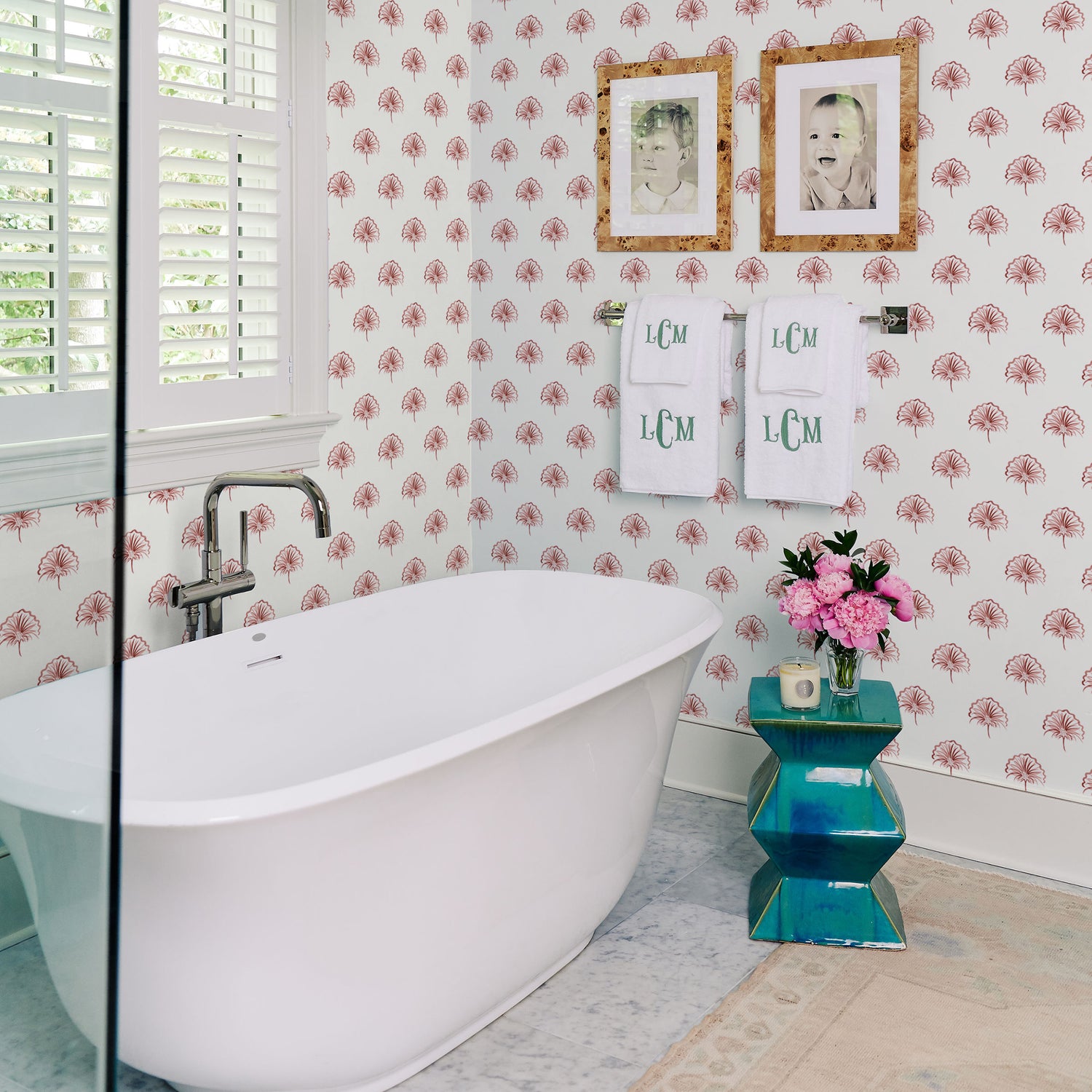 Bathroom styled with Rose Floral printed wallpaper with white bath tub and two photo frames hanging from wall on top of the hanging monogrammed towels