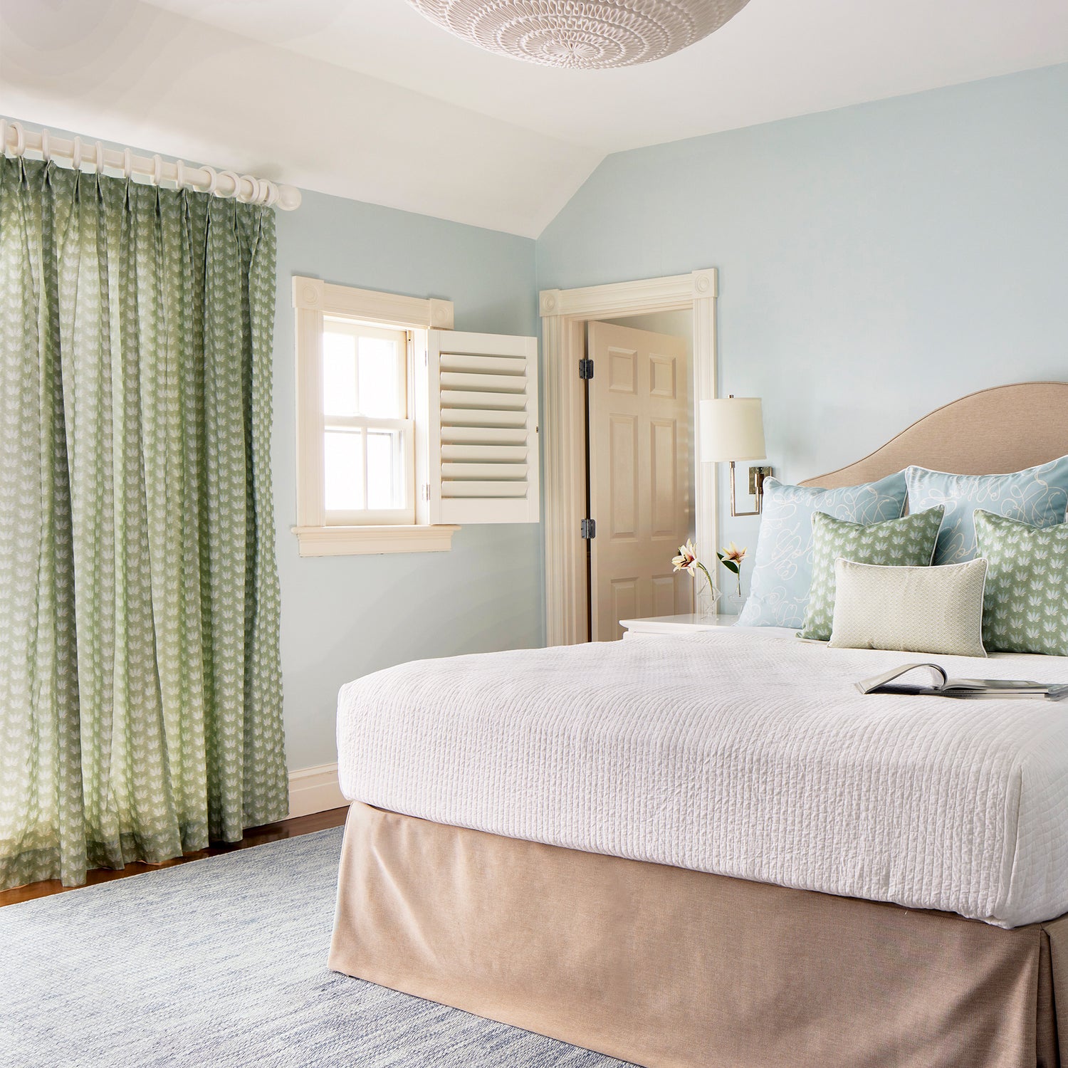 Bedroom with white bed styled with three Powder Blue Abstract Printed Pillows, two Coastal Inspired Green Printed Pillows, and one Moss Green Geometric Printed Lumbar laying by stacked magazines and Blue & Green Floral Drop Repeat Printed Curtains on illuminated window