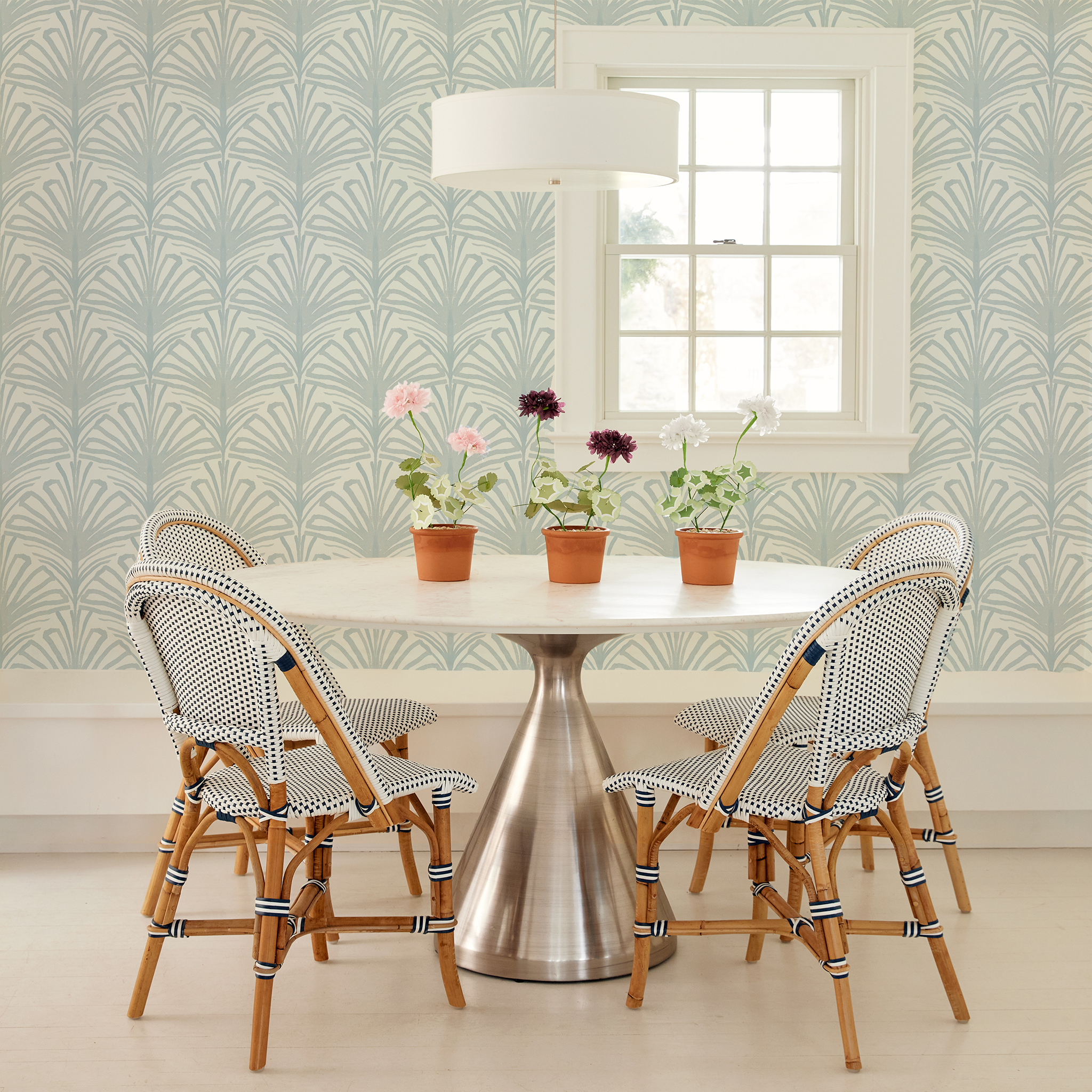 Circular table with flowers on three clay pots with 4 wooden chairs around it next to an illuminated window styled with a Sky Blue Palm Printed Wallpaper